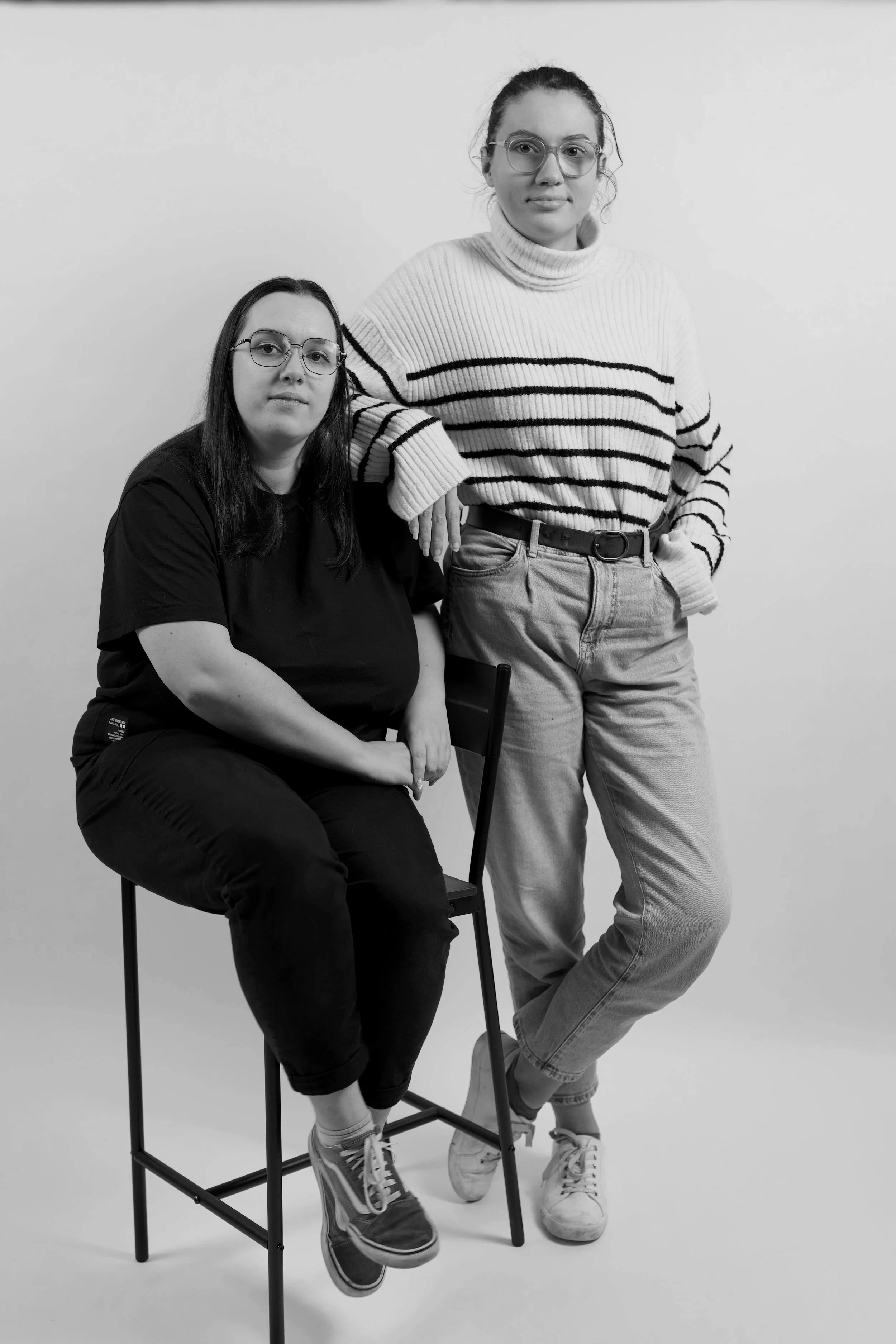 Deux femmes portant des lunettes posent contre un mur blanc. L'une est assise sur une chaise et l'autre se tient à côté d'elle, appuyée sur la chaise. Elles portent des vêtements décontractés.