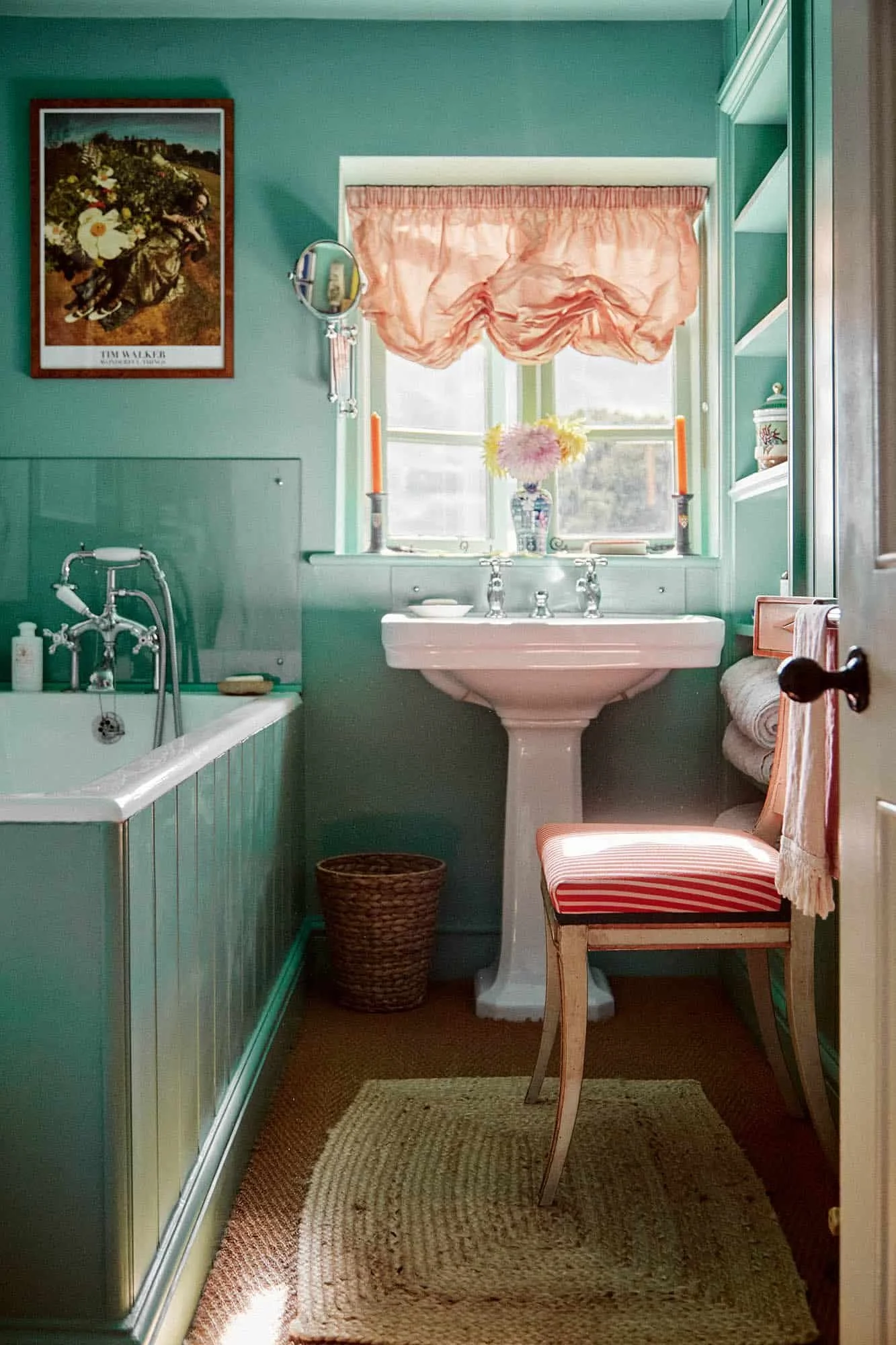 A vintage-style bathroom with teal walls, featuring a window with pink curtains, a white pedestal sink with a vase of flowers, a chair with red and white striped upholstery, a wicker basket, a side cabinet with a towel, and framed artwork on the wall.