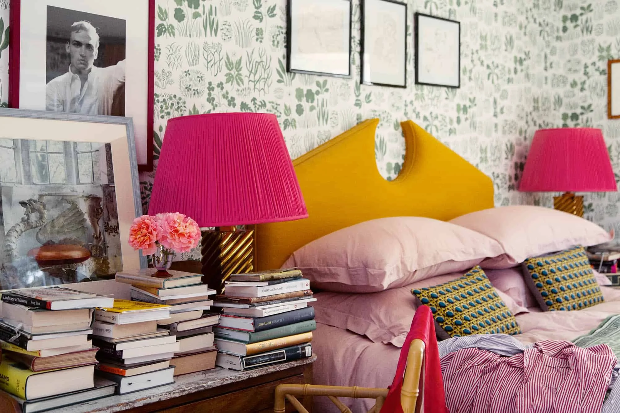 A bedroom with a yellow headboard, pink bedding, two pink lamps, a stack of books on a desk, framed photographs and artwork, and patterned pillows, with a floral wallpaper background.