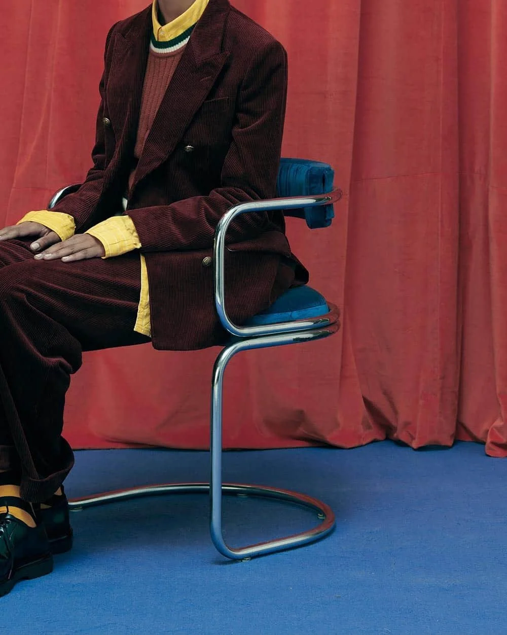 A person wearing a maroon corduroy suit, yellow sweater, and sneakers sitting on a modern blue and chrome chair in front of a pink curtain.