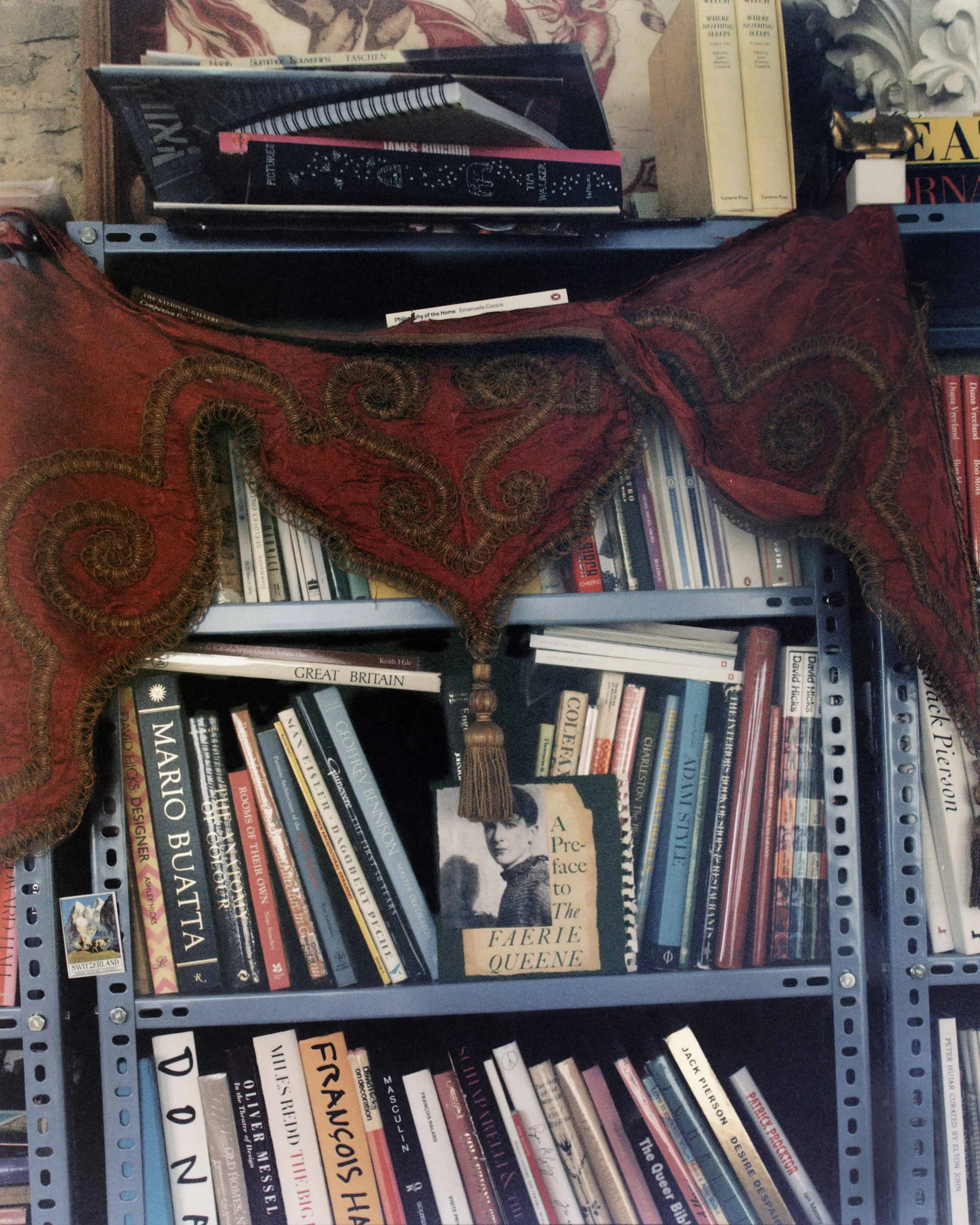 Books on a shelf, with some stacked horizontally and others vertically. The titles include 'Great Britain,' 'Marie Antoinette,' and 'A Pre-face to The Faerie Queene'. Some books are partially obscured by a red decorative fabric with intricate embroidery draped over the shelf.