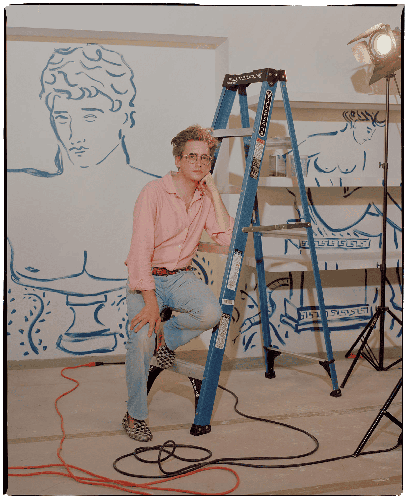 A person sitting on a black chair with a playful pose in front of a white wall with blue line art of classical sculptures, a stepladder, and studio lighting equipment.