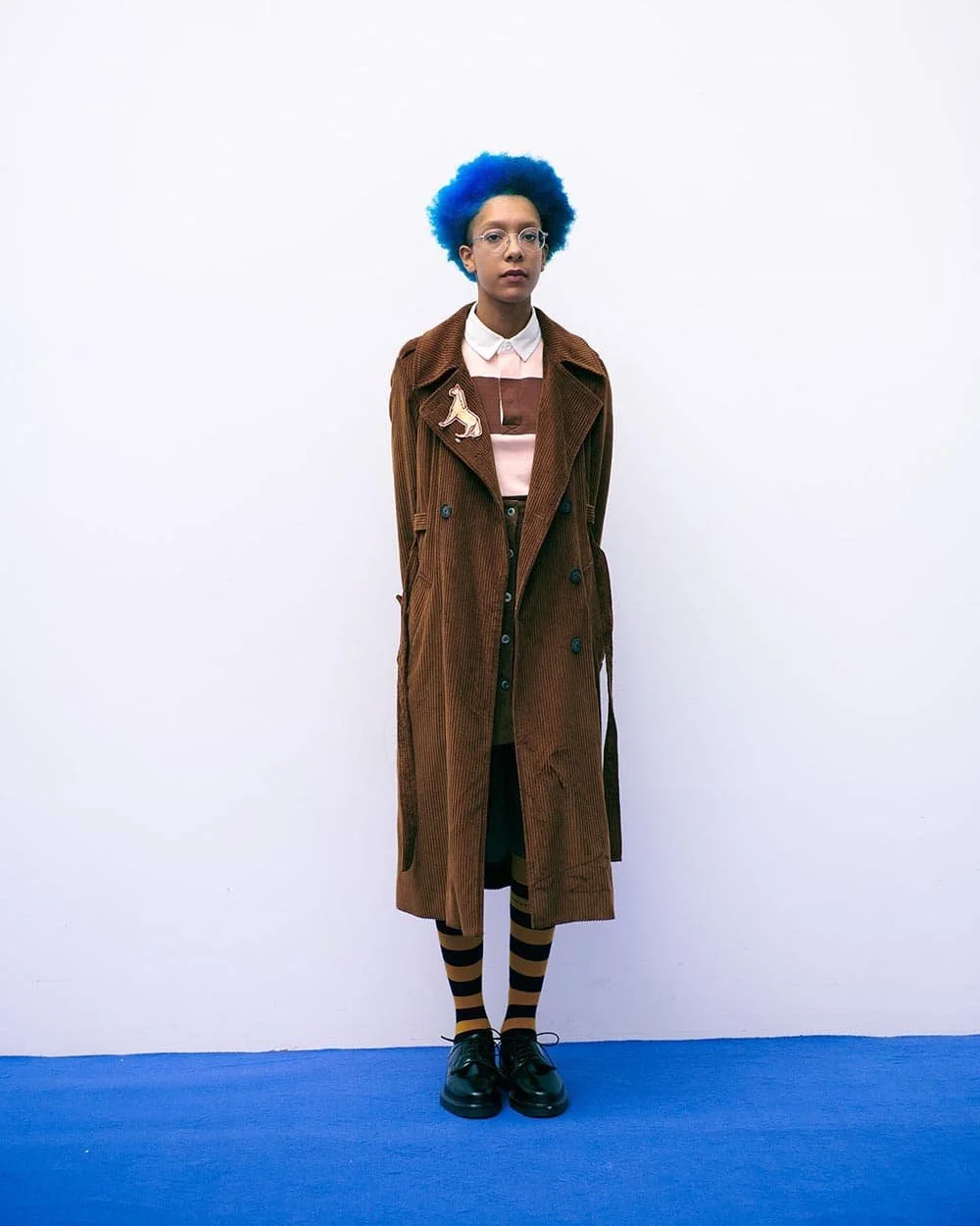 A person with blue curly hair wearing glasses, a long brown coat, a pink and white top, striped socks, black shoes, and standing against a plain white wall.