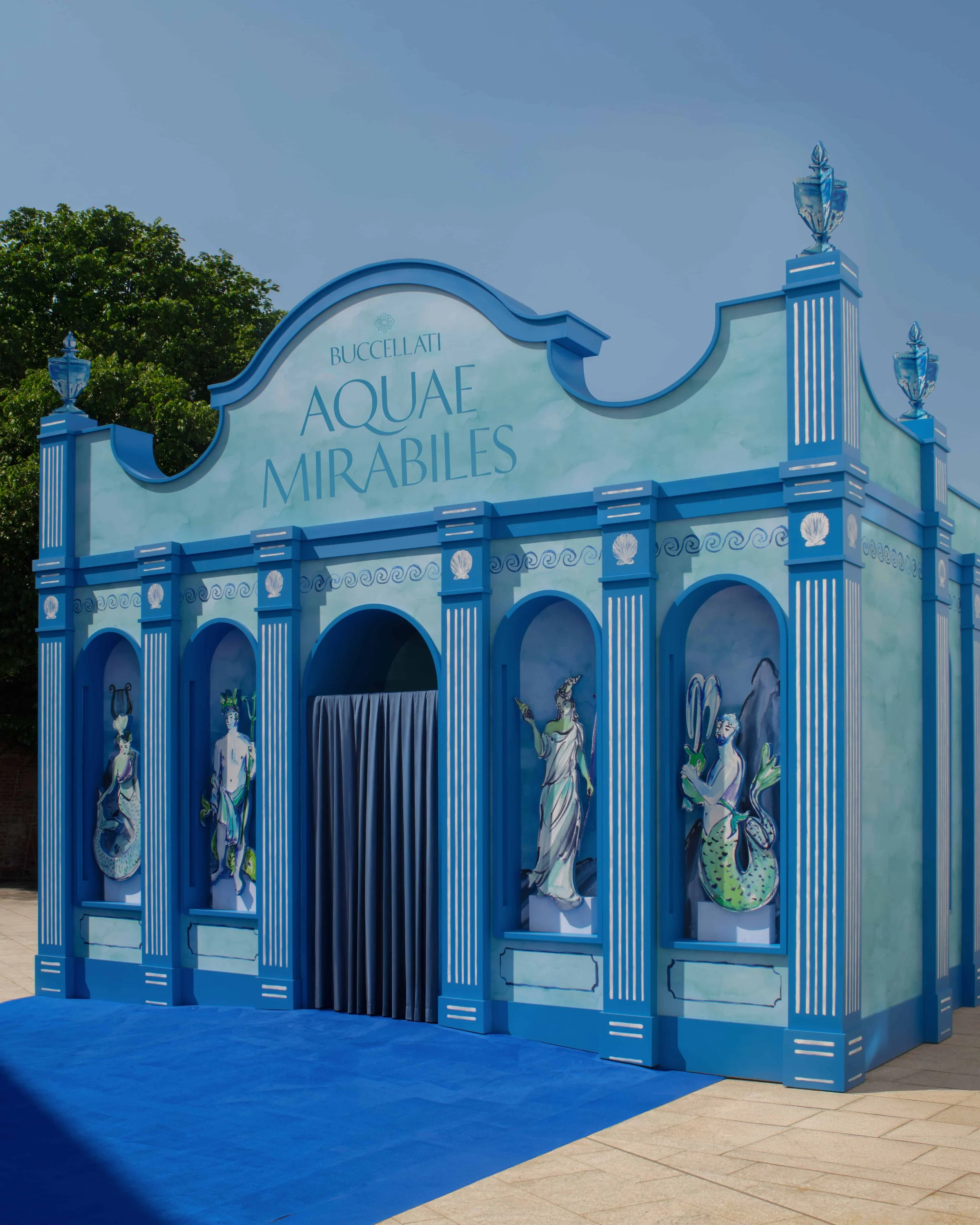 Entrance decorative arch with the words "Buccellati Aquae Mirabiles" in blue, flanked by painted panels of Greek mythological figures, blue flooring, and a small tile area outside.