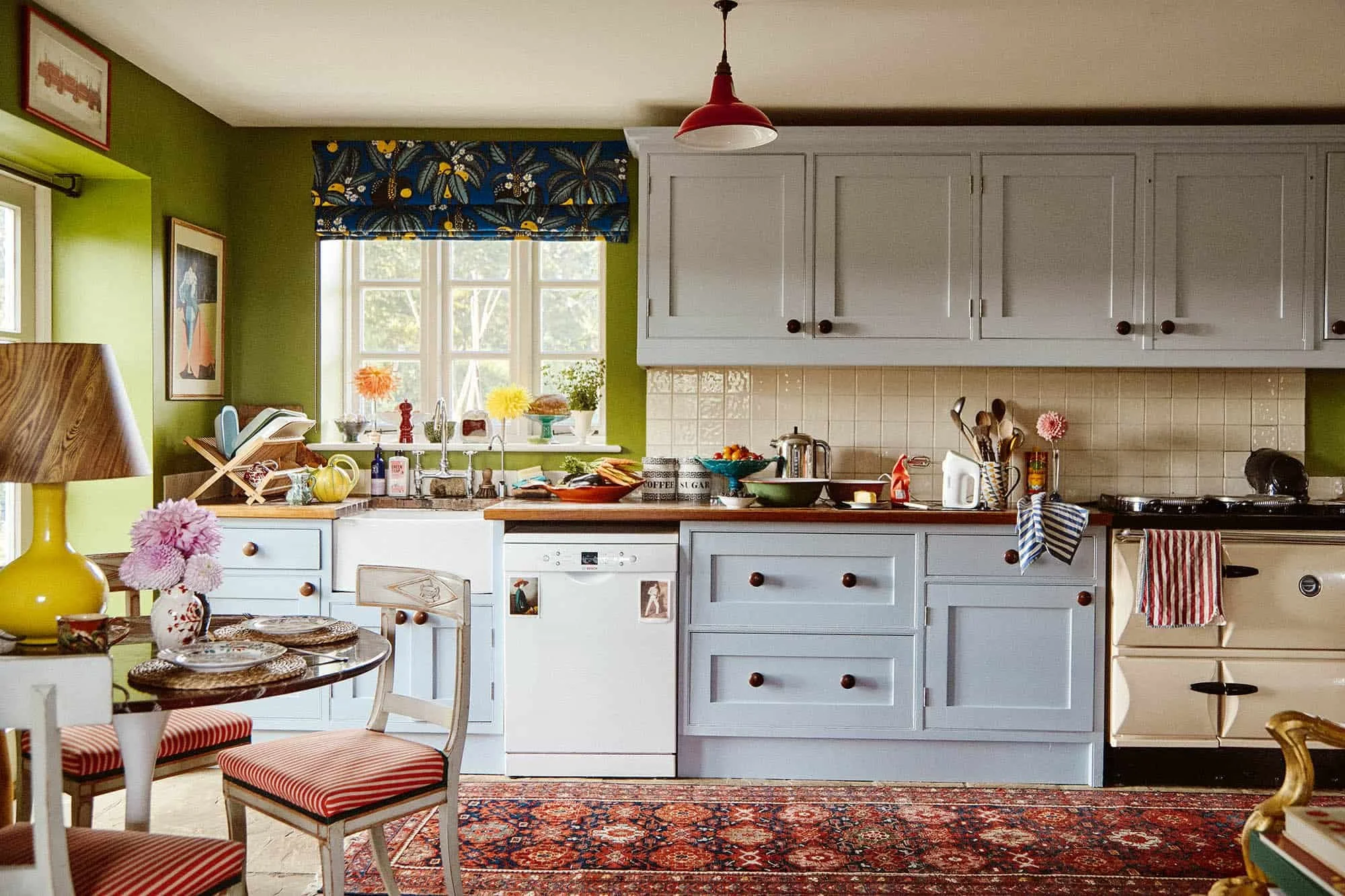 A cozy kitchen with green walls and white cabinets, decorated with colorful flowers and houseplants on the windowsill. There's a small dining table with red and white striped chairs, and various kitchen appliances and utensils on the countertop, including a microwave, toaster, and kettle. A patterned area rug covers the floor.