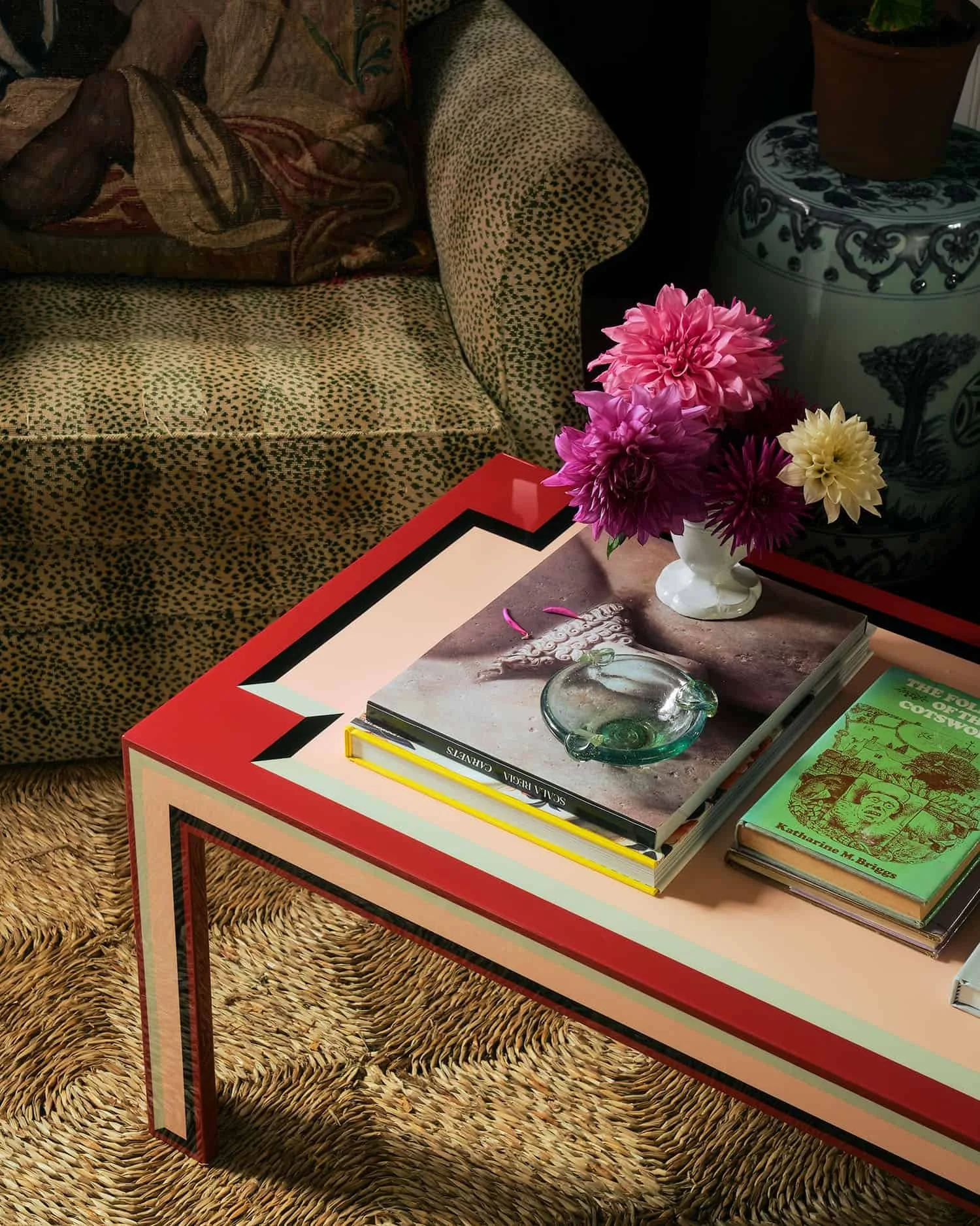 A colorful coffee table with a pink border, topped with a white vase containing pink, purple, yellow, and dark red flowers, several books including 'Solía ser una niña' and 'Katherine M. Briggs,' and a small green book, in a room with a leopard-print armchair and a woven rug.