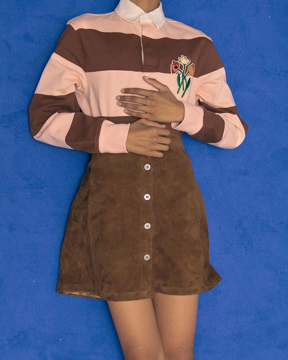 Person lying on a blue surface, wearing a striped pink and brown long-sleeve shirt with flower embroidery on the chest, a brown button-front skirt, and beige stockings.