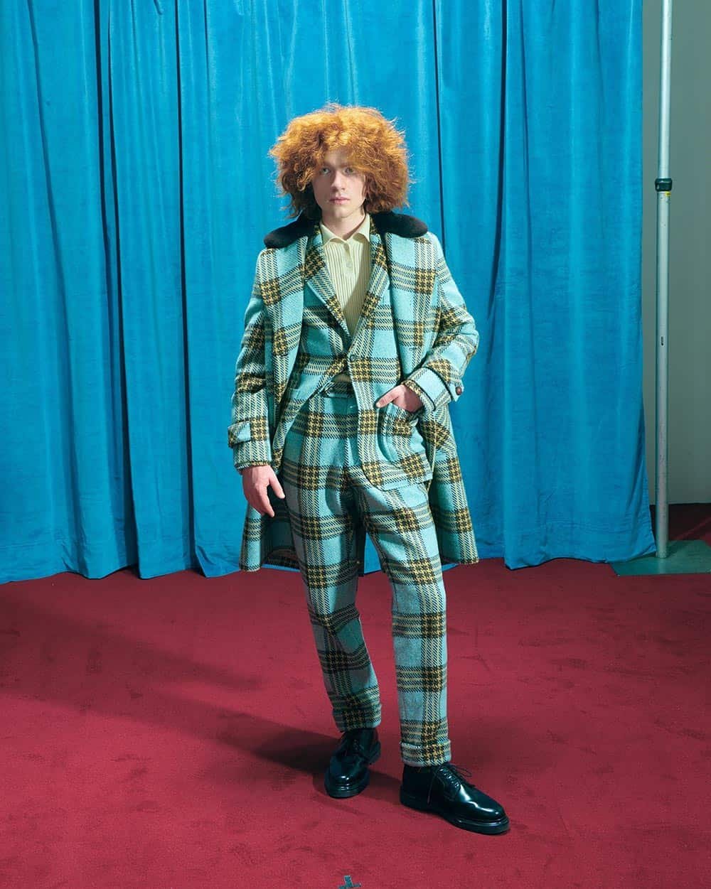 Person with curly red hair wearing a matching plaid suit with a long coat, standing on a red carpet in front of blue curtains.