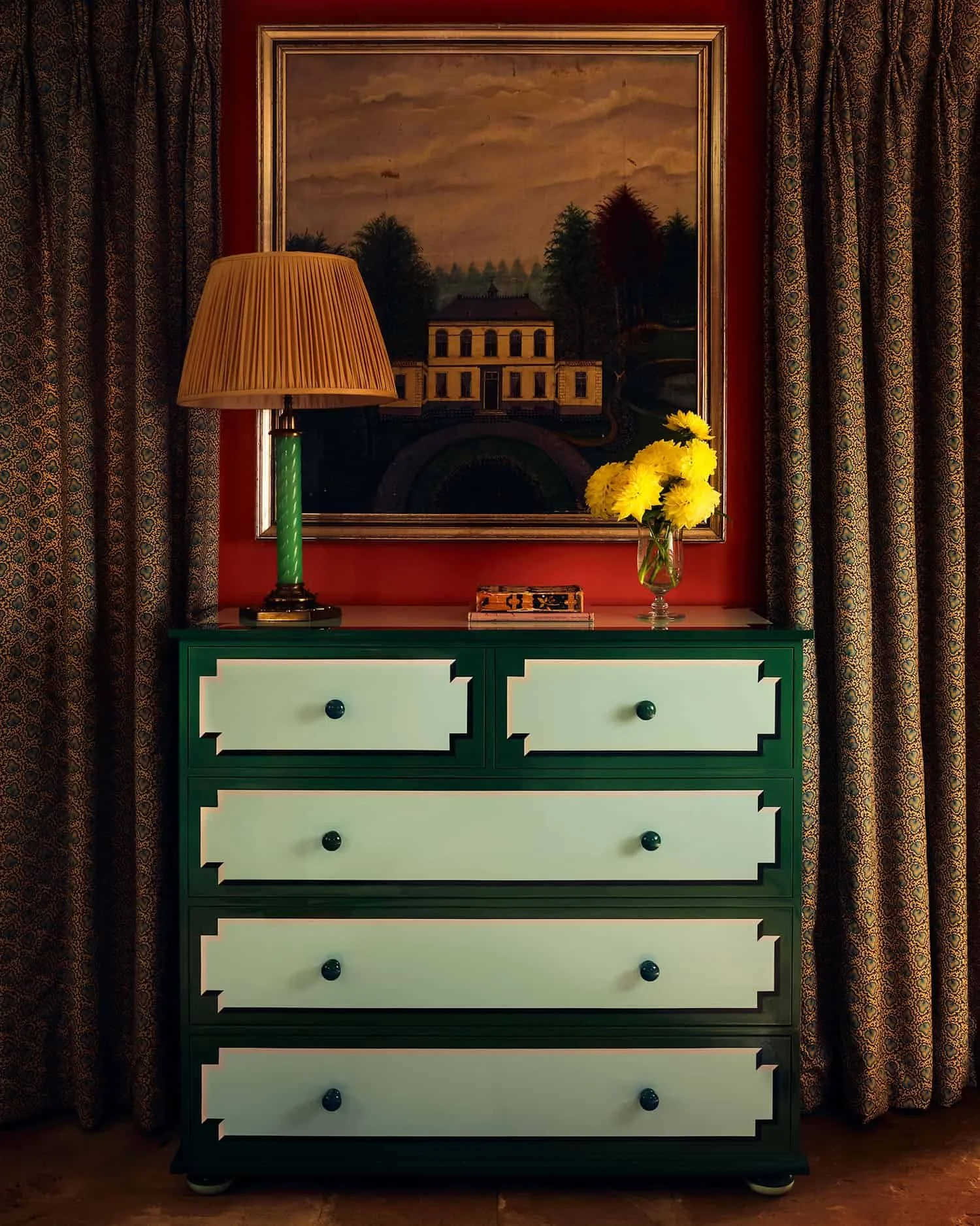 A teal and white dresser with five drawers and black knobs, topped with a yellow flower bouquet in a glass vase, a wooden box, a green table lamp, a framed landscape painting, and patterned curtains on either side.