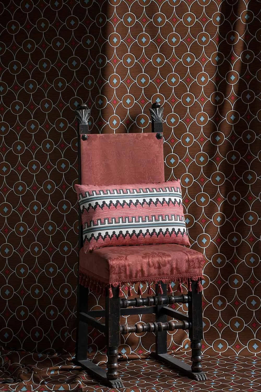 A vintage wooden chair with a red upholstered seat and backrest, decorated with tassels, set against a patterned wallpaper with a geometric design of circles and diamonds in red, white, and blue.