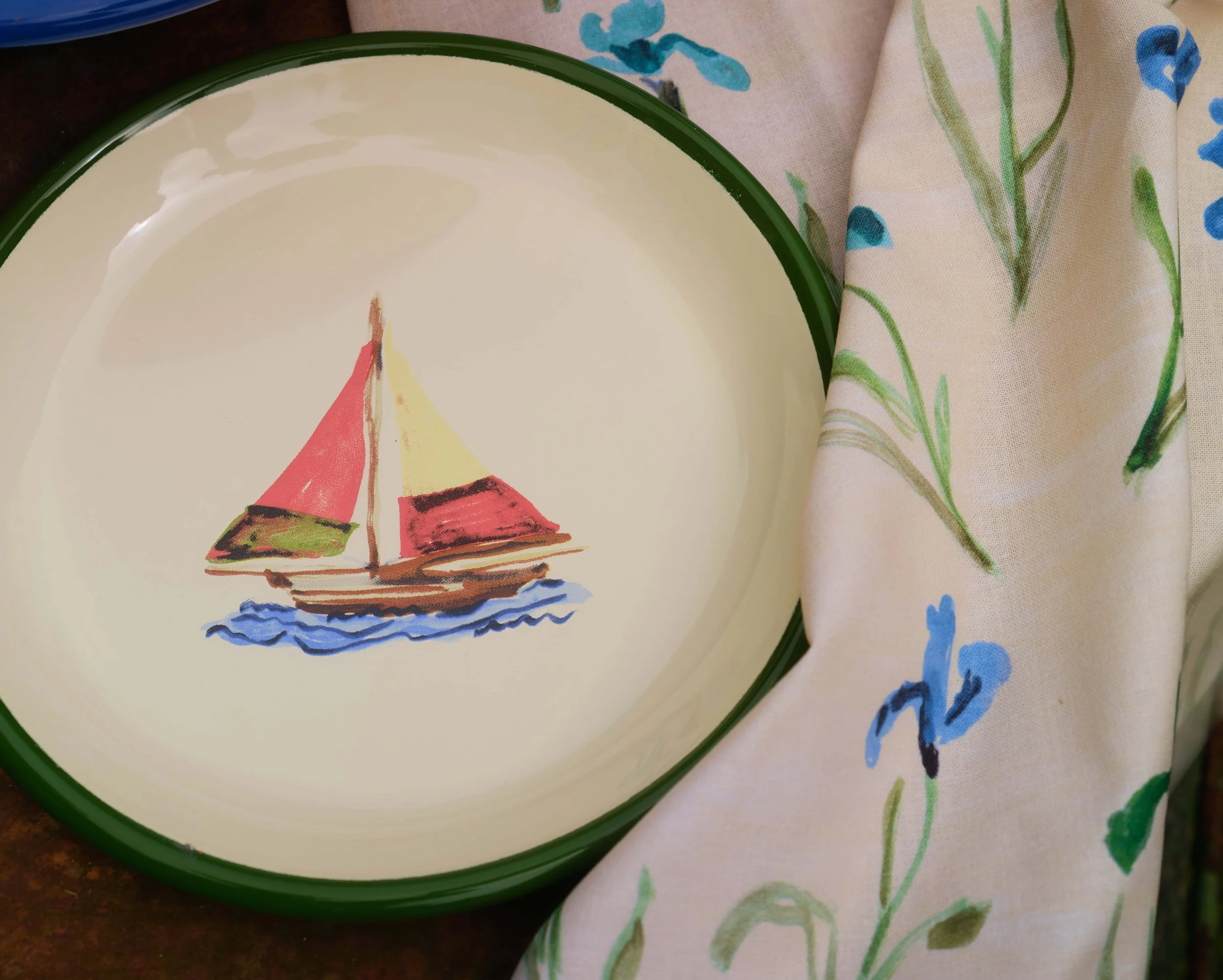 A close-up of a round plate with a green border, featuring a simple illustration of two sailboats on water, placed on a fabric with a floral and leafy pattern.