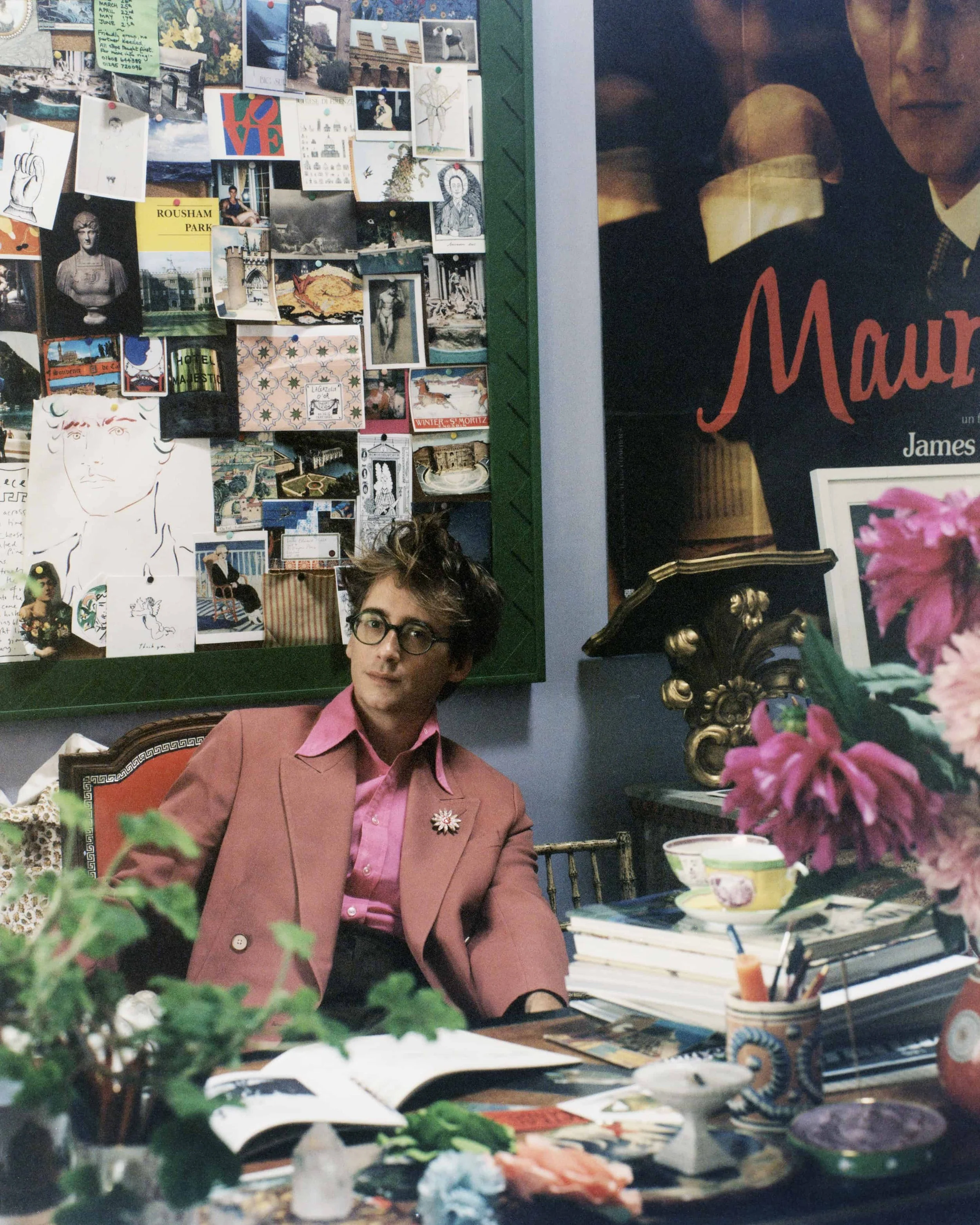 A person with glasses and dark hair, wearing a pink blazer and pink shirt, sitting at a table with stacked papers, books, and art supplies, with a cluttered background of photographs, postcards, and artwork on a wall, and a large poster partially visible on the right.