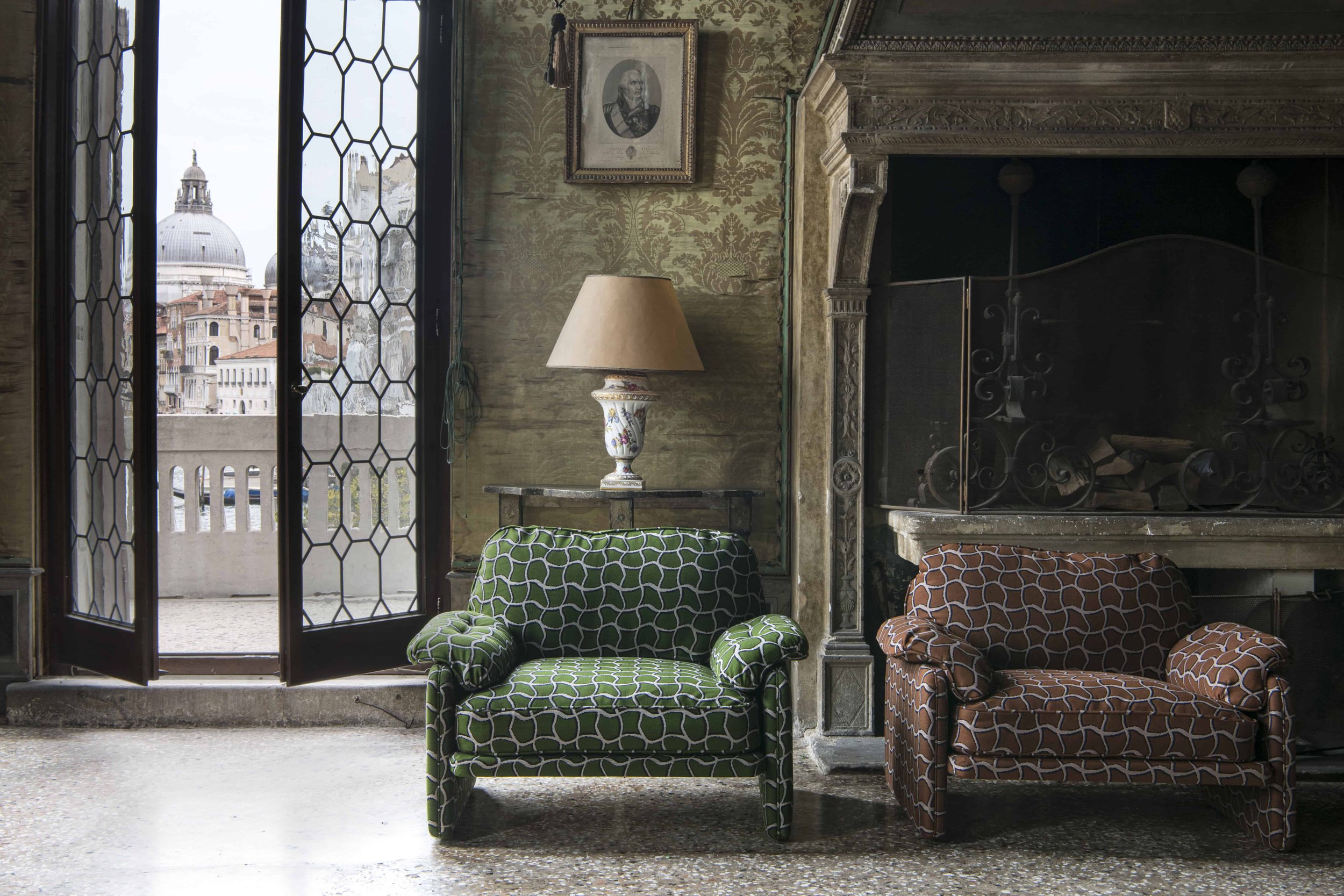 A room with patterned armchairs, a table lamp, a fireplace, and a balcony with a view of historic buildings and a dome.