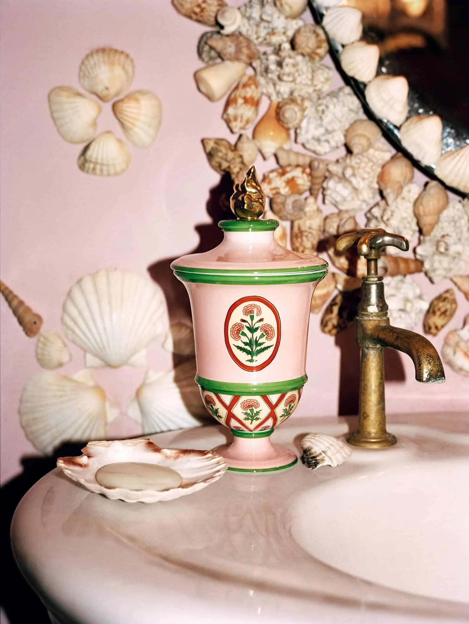 A pink ceramic perfume bottle with green accents and floral patterns, topped with a gold decorative lid, next to a copper faucet on a pink surface, with seashells and a pink wall decorated with seashells in the background.
