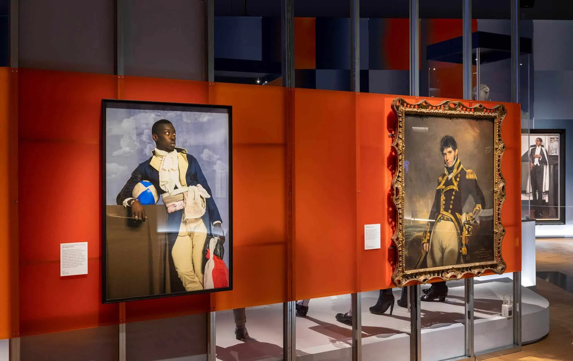 Museum exhibit showcasing portraits and a historical painting of a man in a dark military uniform with gold accents, alongside a framed modern portrait of a person in a navy jacket and holding a football, set against a red background.