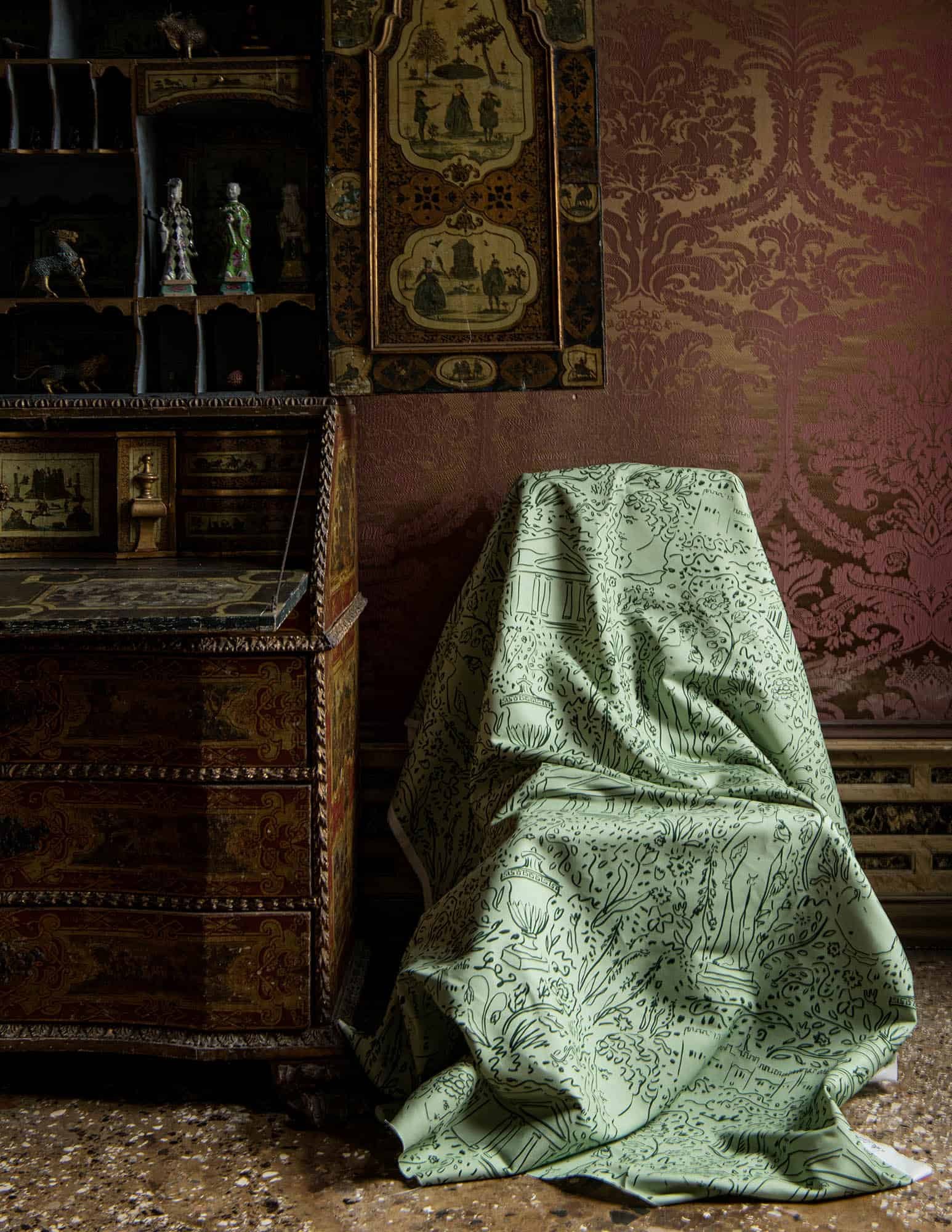 An interior scene with a wooden cabinet on the left, displaying various decorative figurines and animal sculptures. A tapestry with historical or pastoral scenes hangs on the wall behind the cabinet. To the right, a piece of fabric with intricate designs covers a chair or small table, with part of the fabric spilling onto the textured floor. The overall ambiance is vintage and antique.