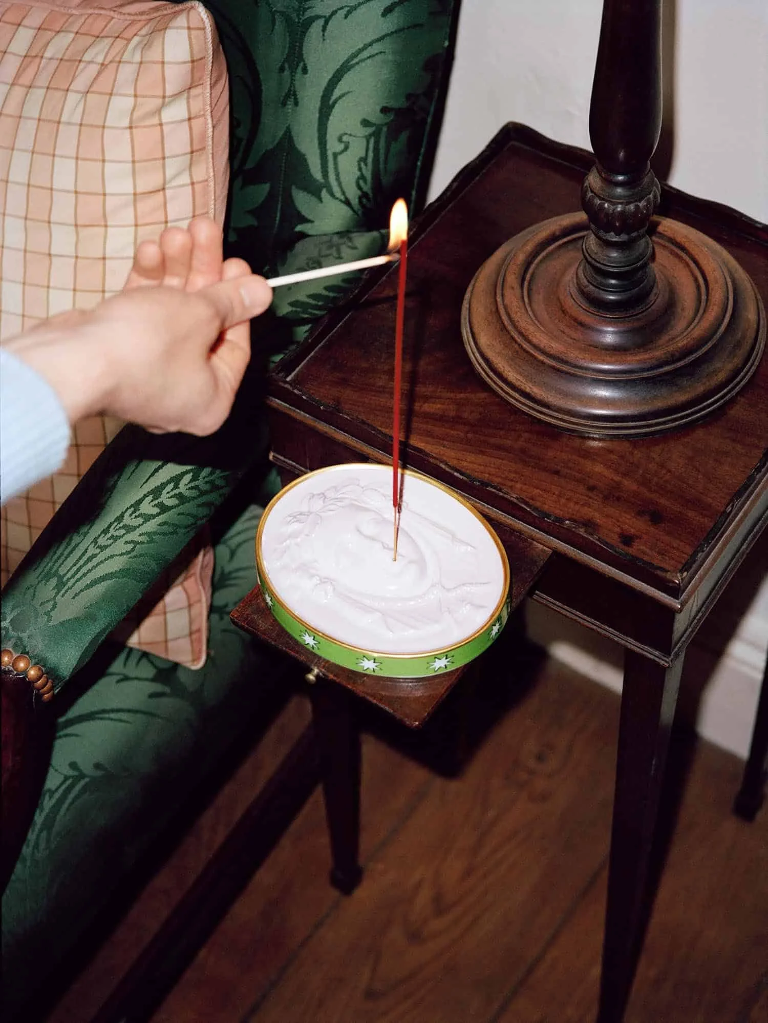 Person lighting a red candle with a match. The candle is in a decorated container, placed on a wooden side table. The person is sitting on a dark green patterned armchair.