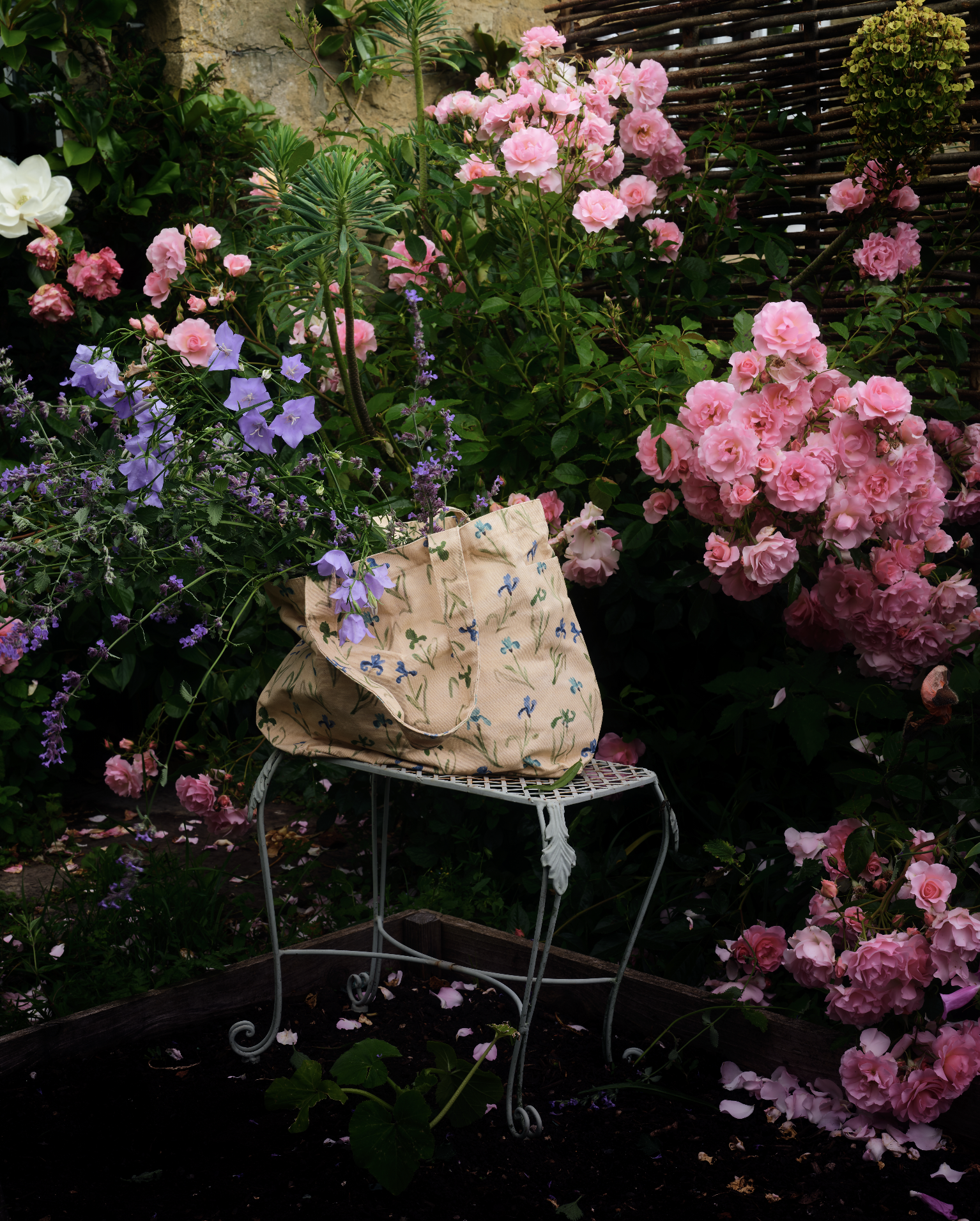 A garden filled with pink and purple flowers, including roses, with a white metal table and a cloth bag on top, surrounded by lush greenery and a wooden fence.