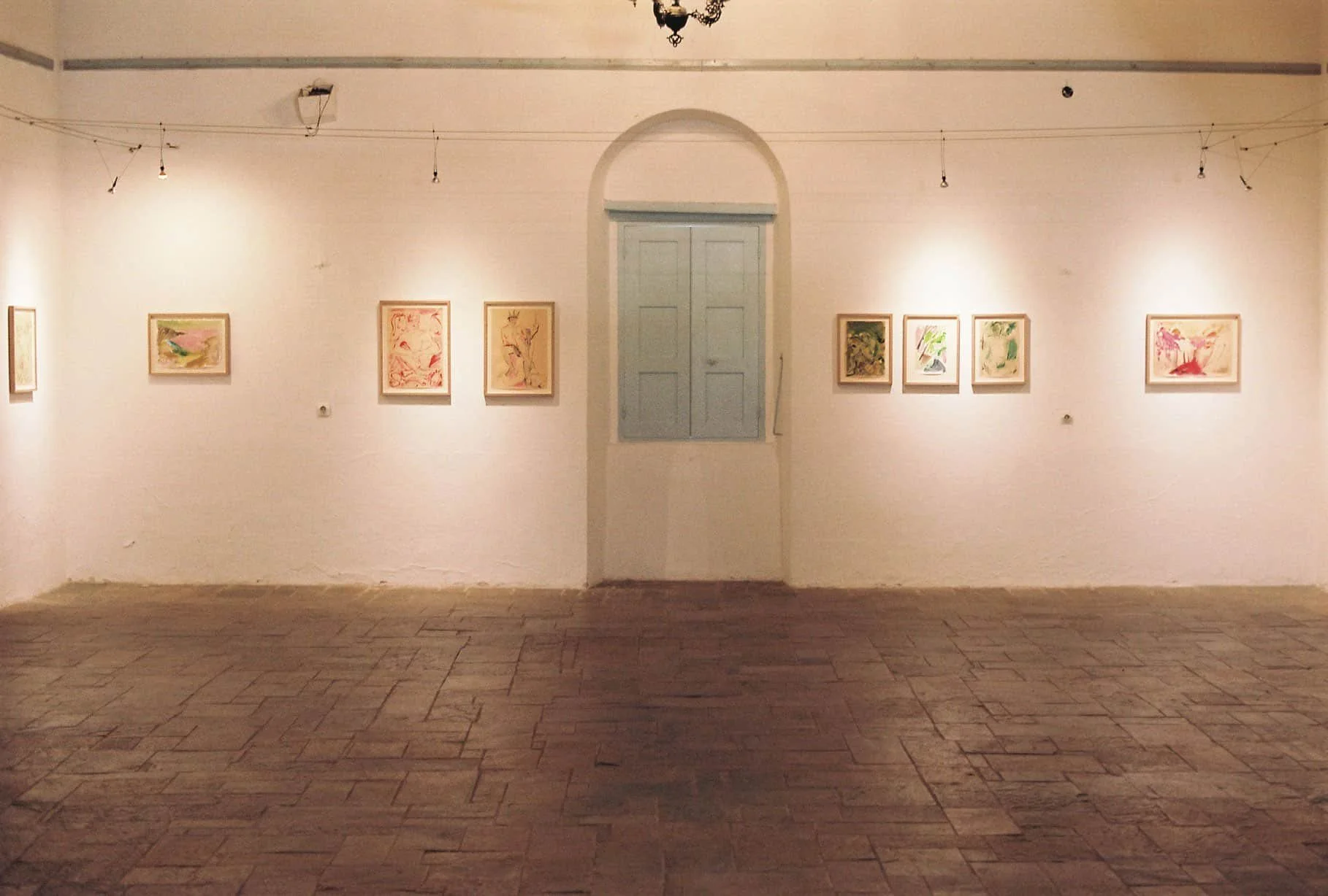 An art gallery with framed artwork on white walls, a closed blue door in the center, wooden floor, and ceiling lights.