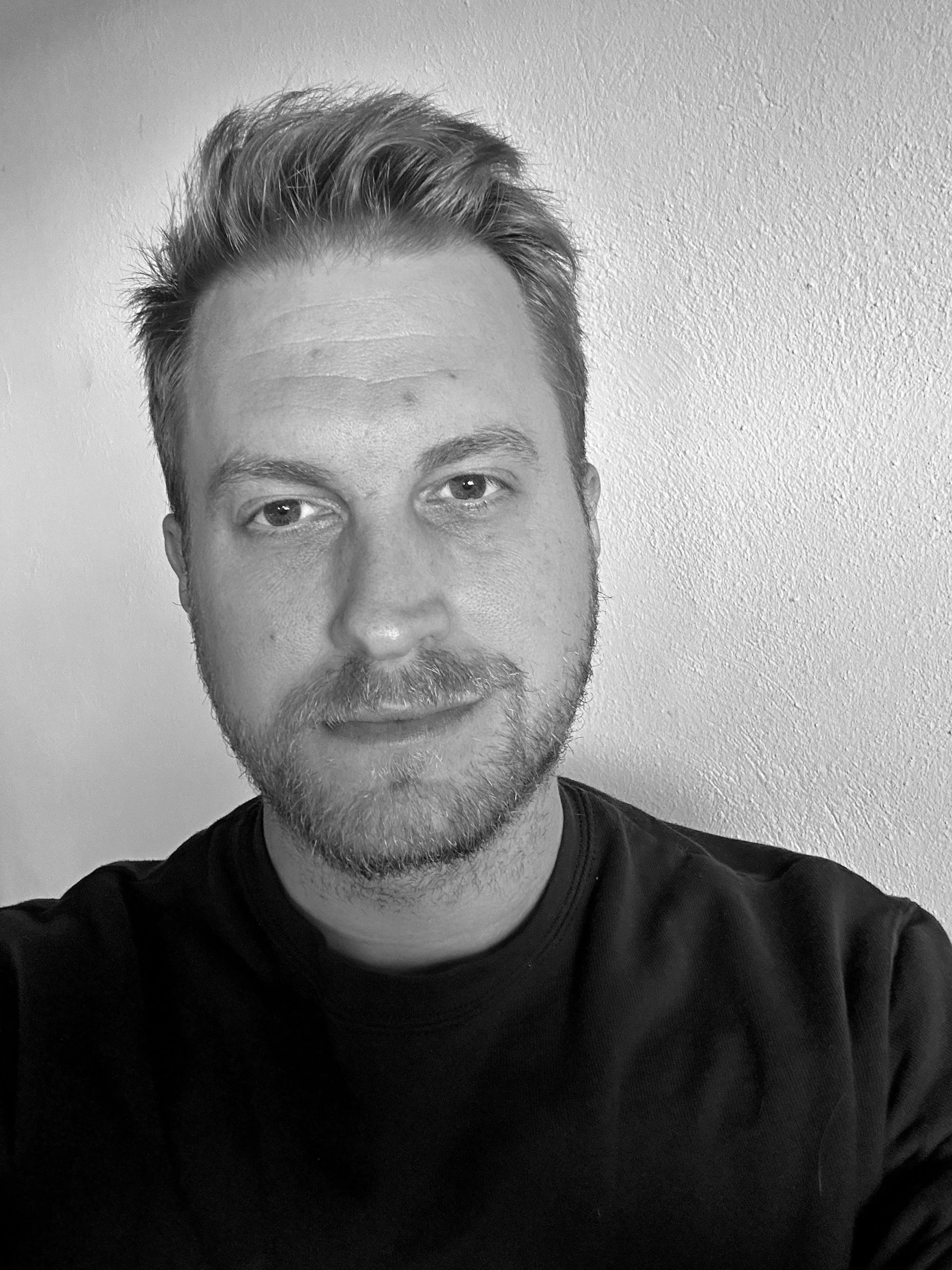 Schwarz-weiß Porträt eines Mannes mit kurzen, gewellten Haaren, Bart und freundlichem Ausdruck, trägt ein schwarzes T-Shirt, steht vor einer neutralen Wand.