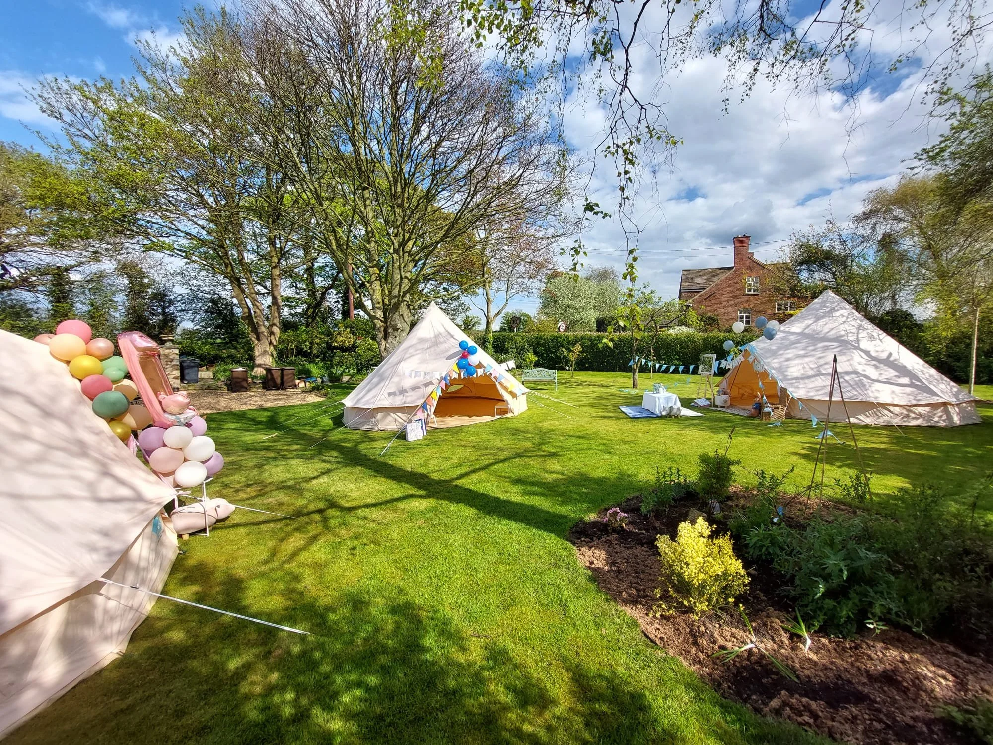 Outdoor garden scene with white tents decorated with balloons and bunting, set on lush green lawn with trees and a brick house in the background on a partly cloudy day.