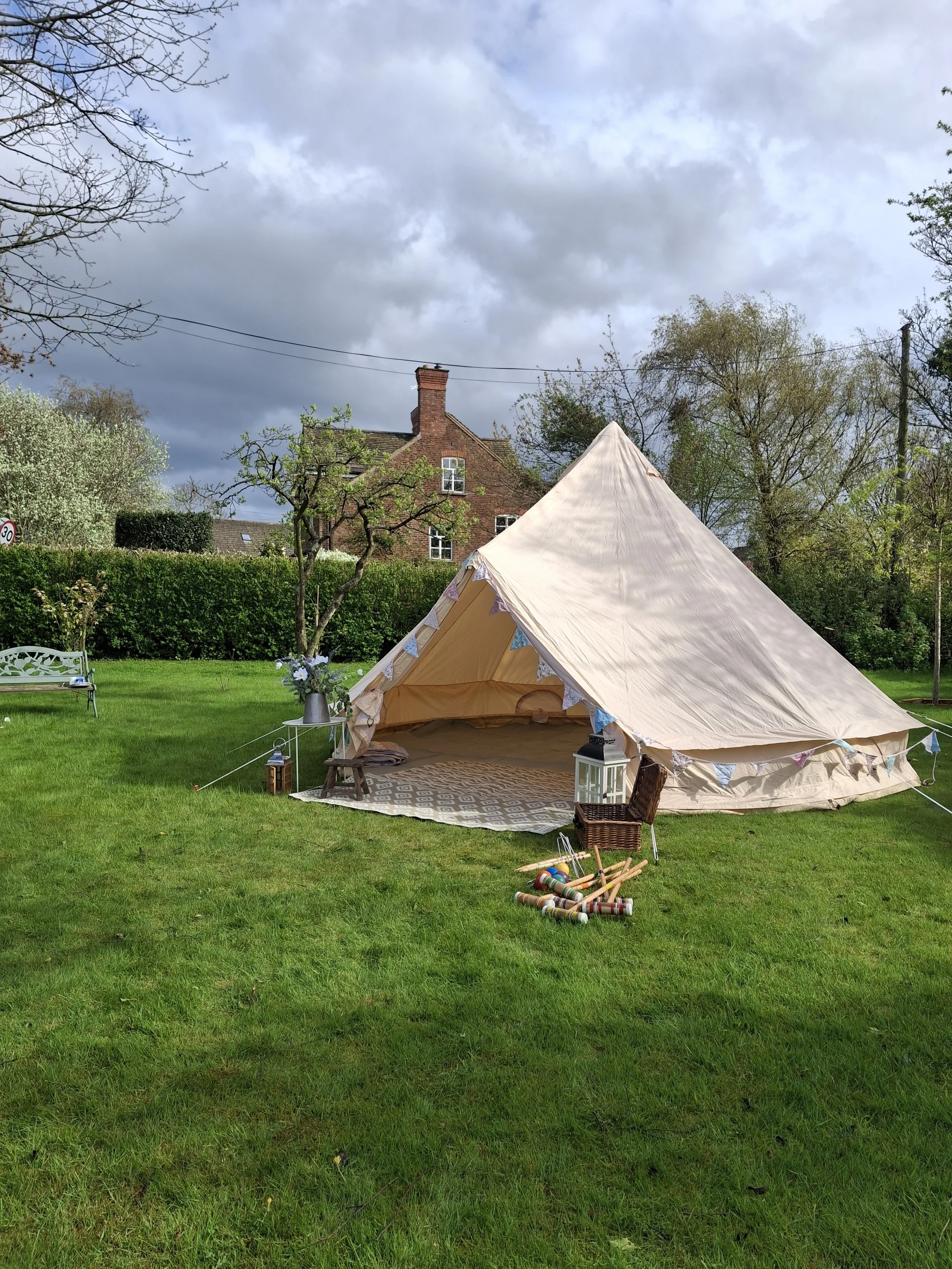 A large cream-colored tent set up on a lush green lawn with a vintage rug underneath. There are small decorative bunting flags hung on the tent. A wicker basket and small wooden chairs are nearby, with a lantern and a umbrella. A flower arrangement in a gray pitcher is placed at the entrance. The background shows a brick house with a chimney, trees with blossoming flowers, and a cloudy sky.