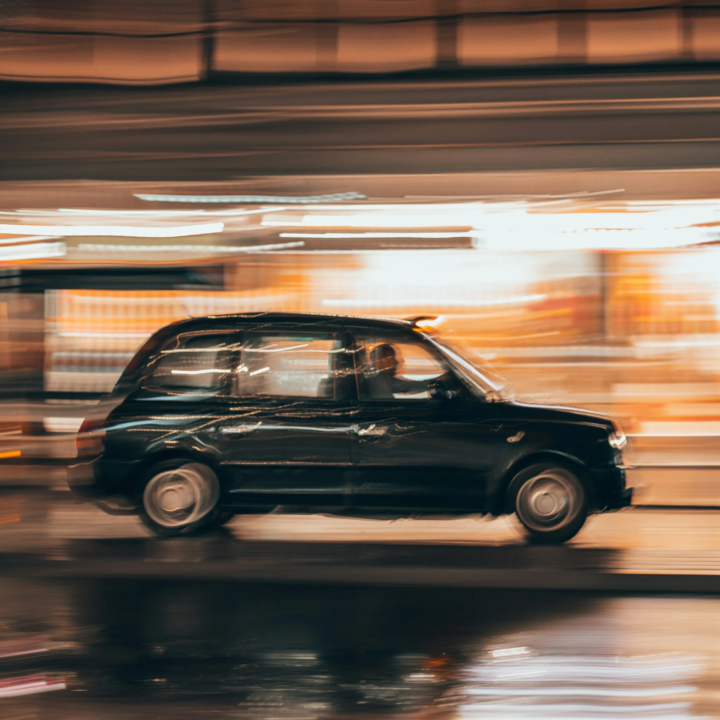 Motion-blurred image of a black car with a sunroof driving through a city at night, with lights reflecting off the wet pavement.