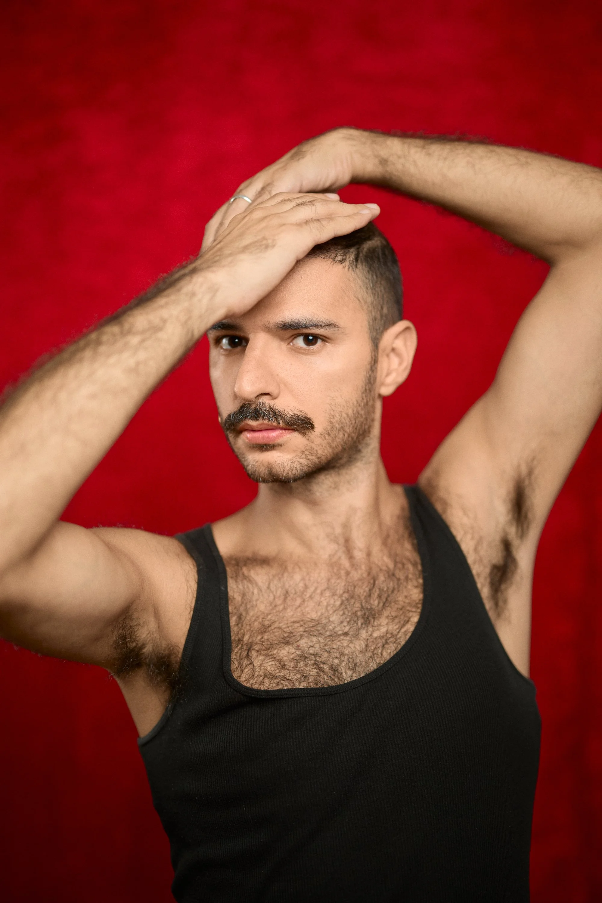A man with dark hair and a mustache posing with his left hand on his head, wearing a black tank top against a red background.