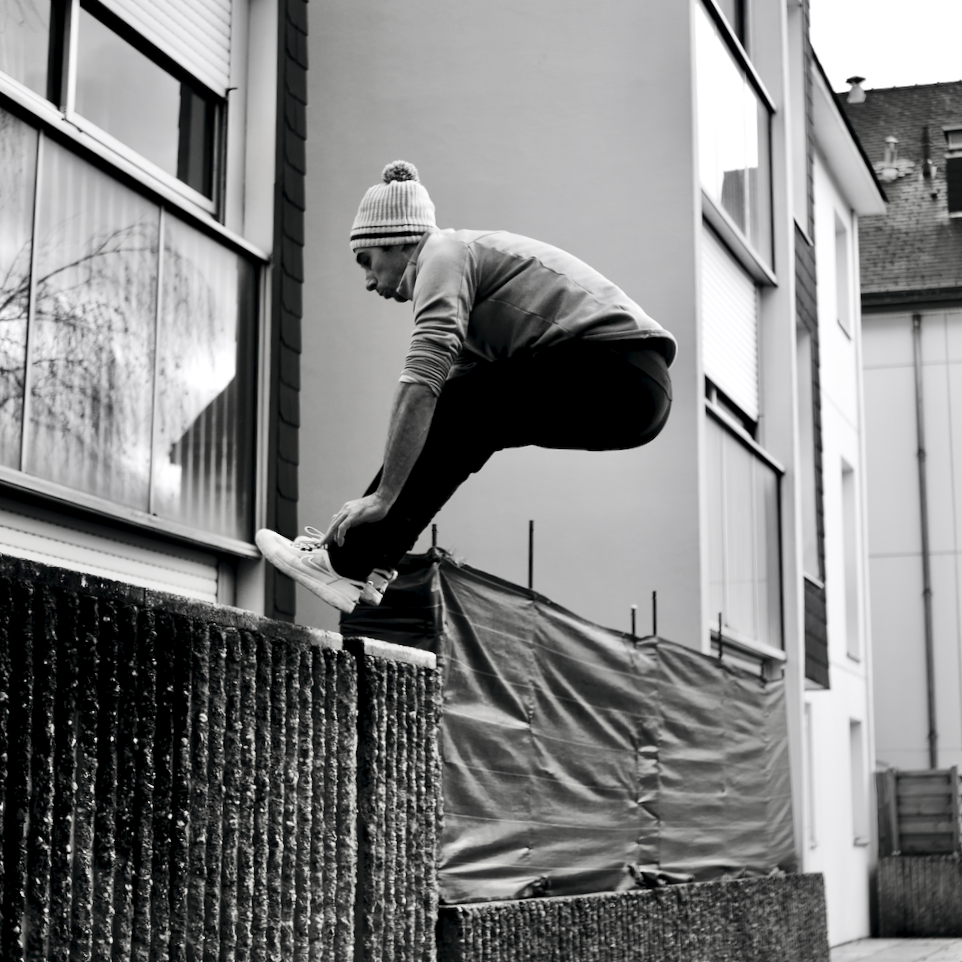Un garçon en sweater et bonnet blanc, en pleine activité de saut ou de saut en parkour devant un bâtiment.