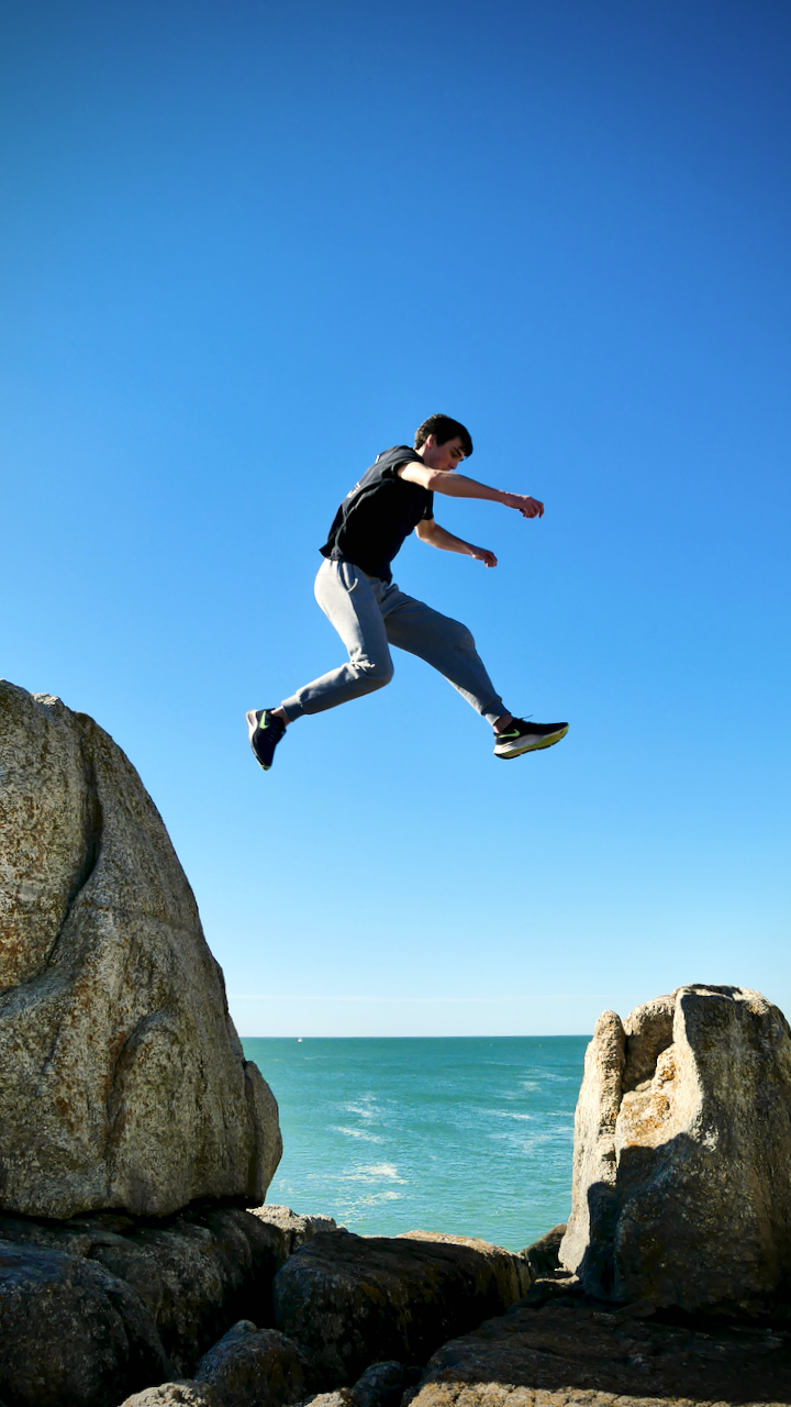 Un jeune homme en plein saut entre deux rochers au bord de la mer, ciel bleu clair en arrière-plan.