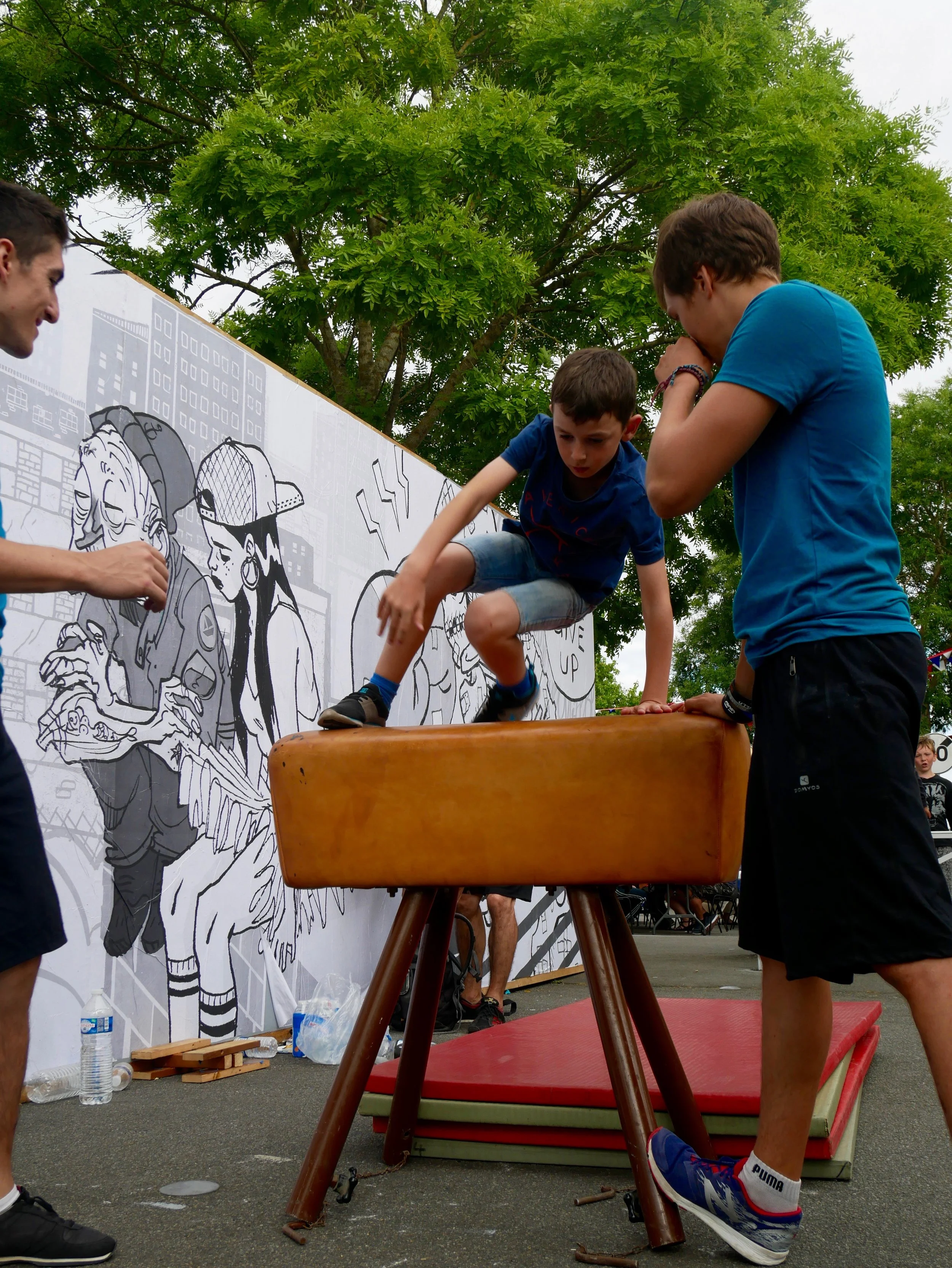 Trois jeunes hommes en train de faire de la gymnastique sur un tapis dans une zone extérieure, avec un mur illustré de dessins de jeunes et de bâtiments en fond, entourés d'arbres verts.
