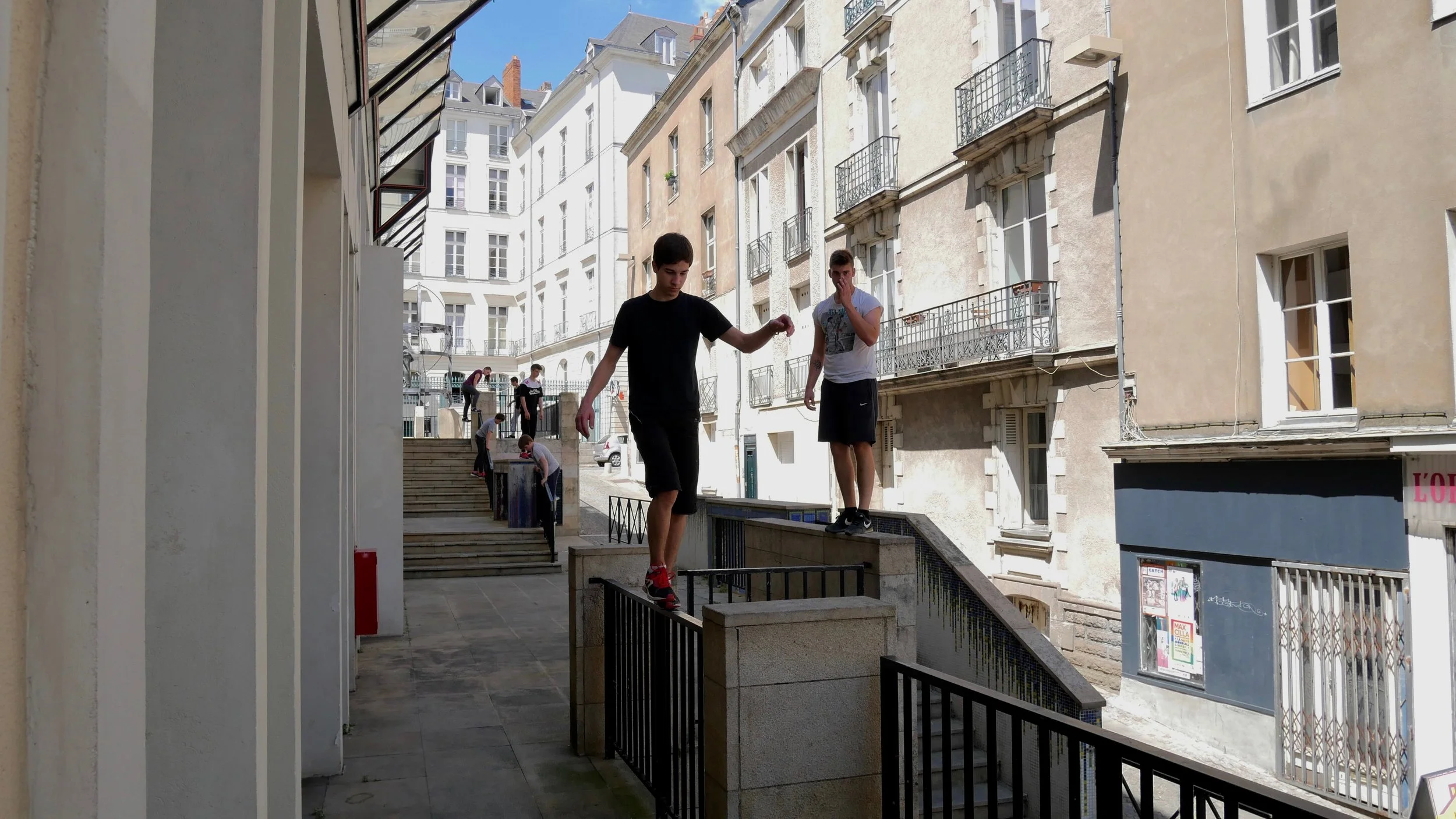 Jeunes pratiquant le parkour à nantes, en ville, avec plusieurs autres personnes en arrière-plan. Il marche en équilibre sur une barrière. stage et cours encadré par un professionnel