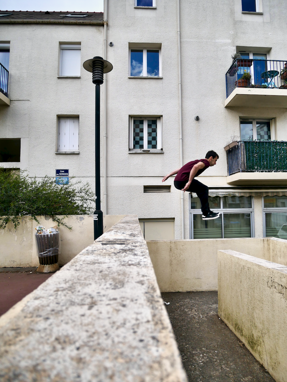 Jeune homme sautant depuis un mur en milieu urbain, avec bâtiments résidentiels en arrière-plan, un lampadaire et une poubelle visible.