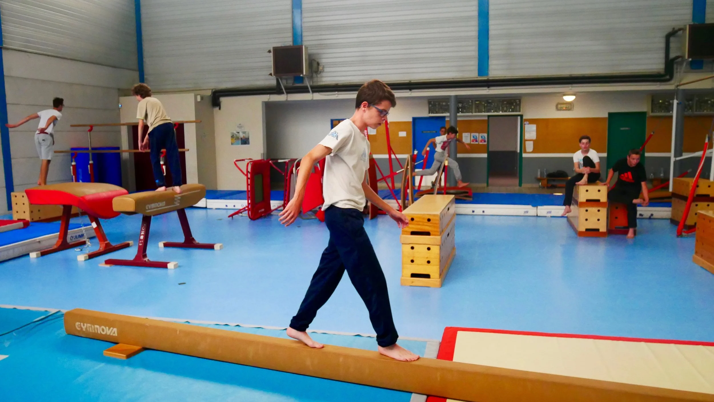 Jeunes pratiquants de gymnastique en salle, sautant et s'entraînant avec des équipements comme des mâts, des tapis et des blocs de gymnastique.