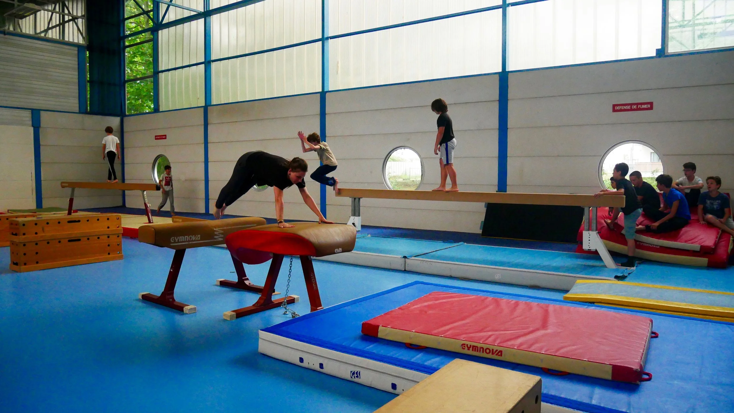 Jeunes pratiquant de la gymnastique dans une salle d'entraînement avec divers appareils, observés par un groupe de personnes en arrière-plan.