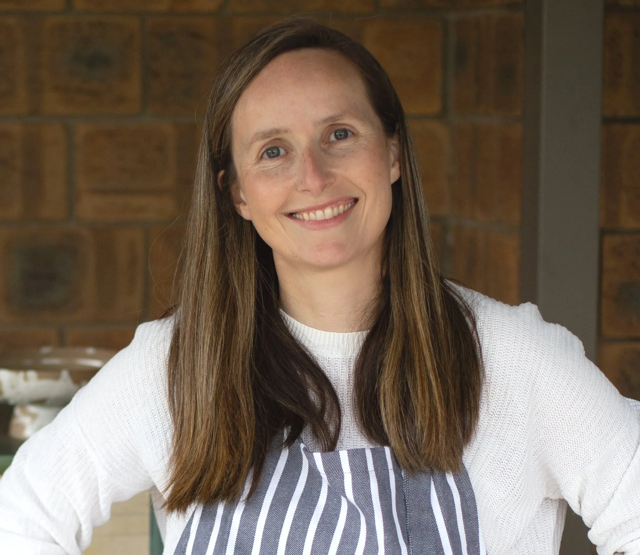 Smiling woman with long brown hair, wearing a white knit sweater and black-and-white striped apron, standing in front of a brick wall.
