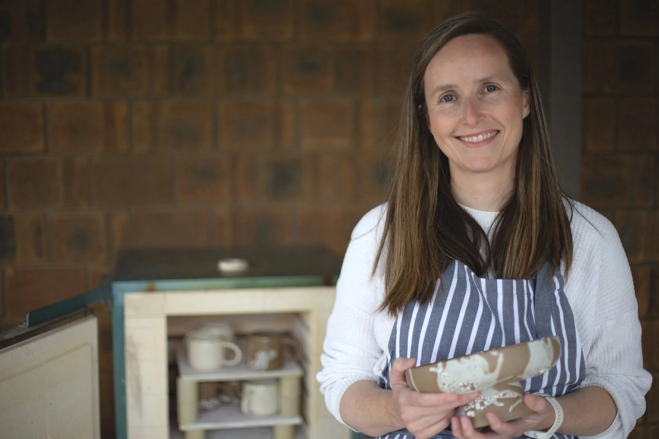 A woman with long brown hair, wearing a white long-sleeve shirt and a navy and white striped apron, smiling and holding a wrapped cake in a bakery or kitchen setting.