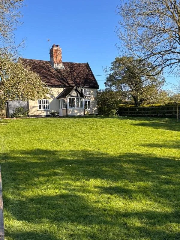 A cozy white house with a red-tiled roof and chimney, surrounded by trees and a well-maintained grassy yard under a clear blue sky.