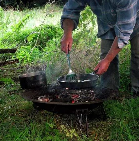 Person cooking on an outdoor grill with two pots and a frying pan in a grassy area.