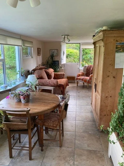 Concervatory with dining area, featuring a round wooden table with four chairs, pink upholstered armchairs, a wooden cabinet, and windows with greenery outside.
