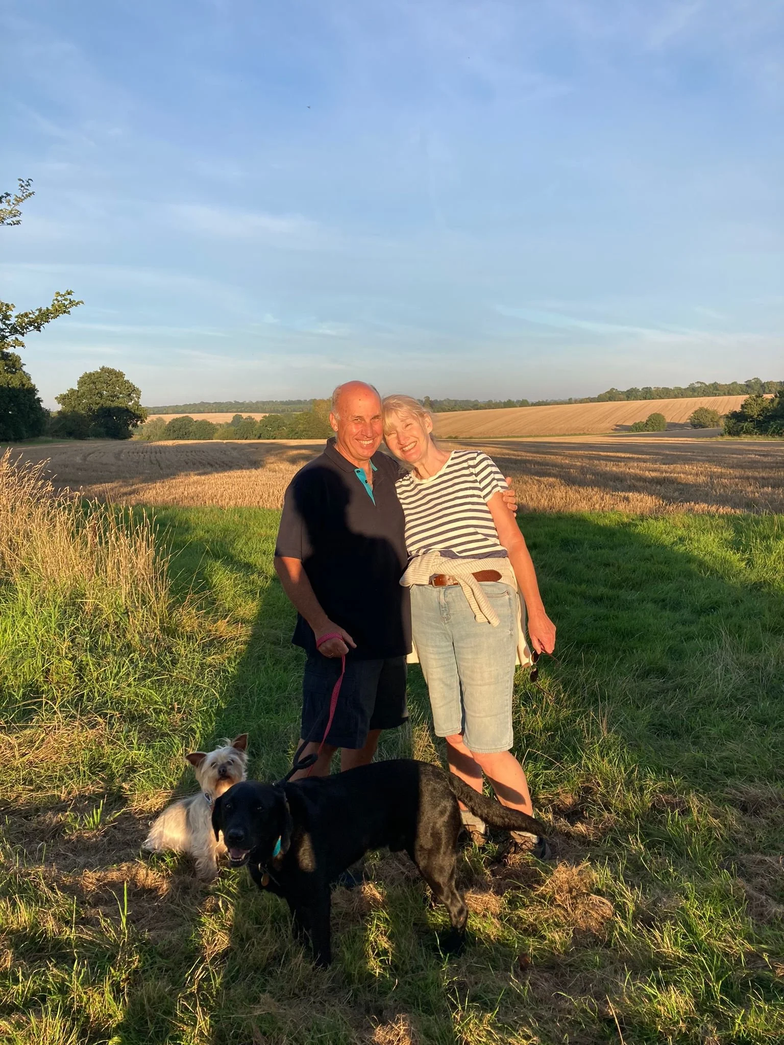 Jane and Nic Pike, smiling  and  hugging each other outdoors in a field with two dogs at their feet, with a blue sky and rolling countryside in the background.