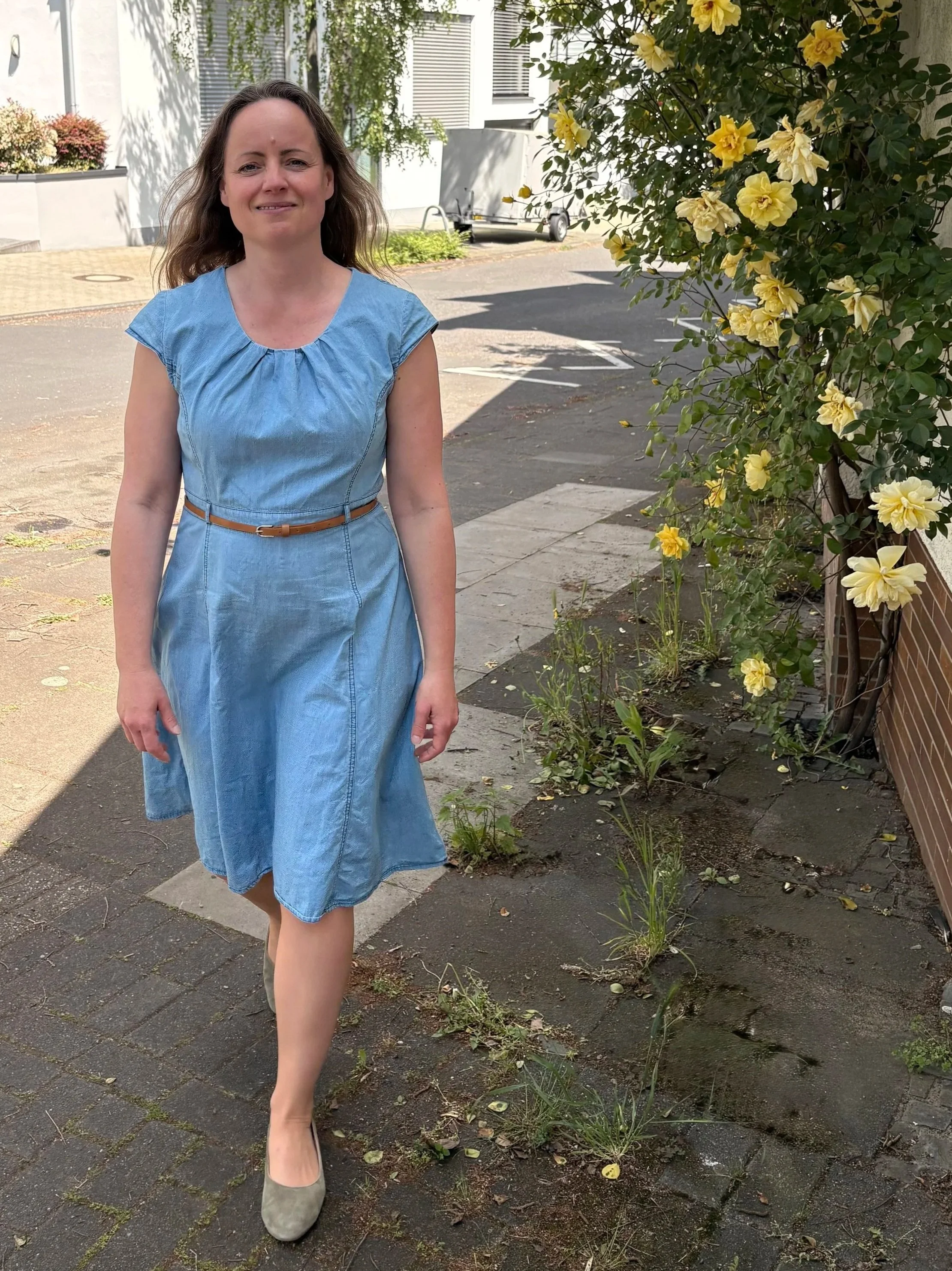 Eine Frau in einem blauen Kleid läuft auf einer Steinterrasse vorbei an einem Blumenbeet mit gelben Blumen.