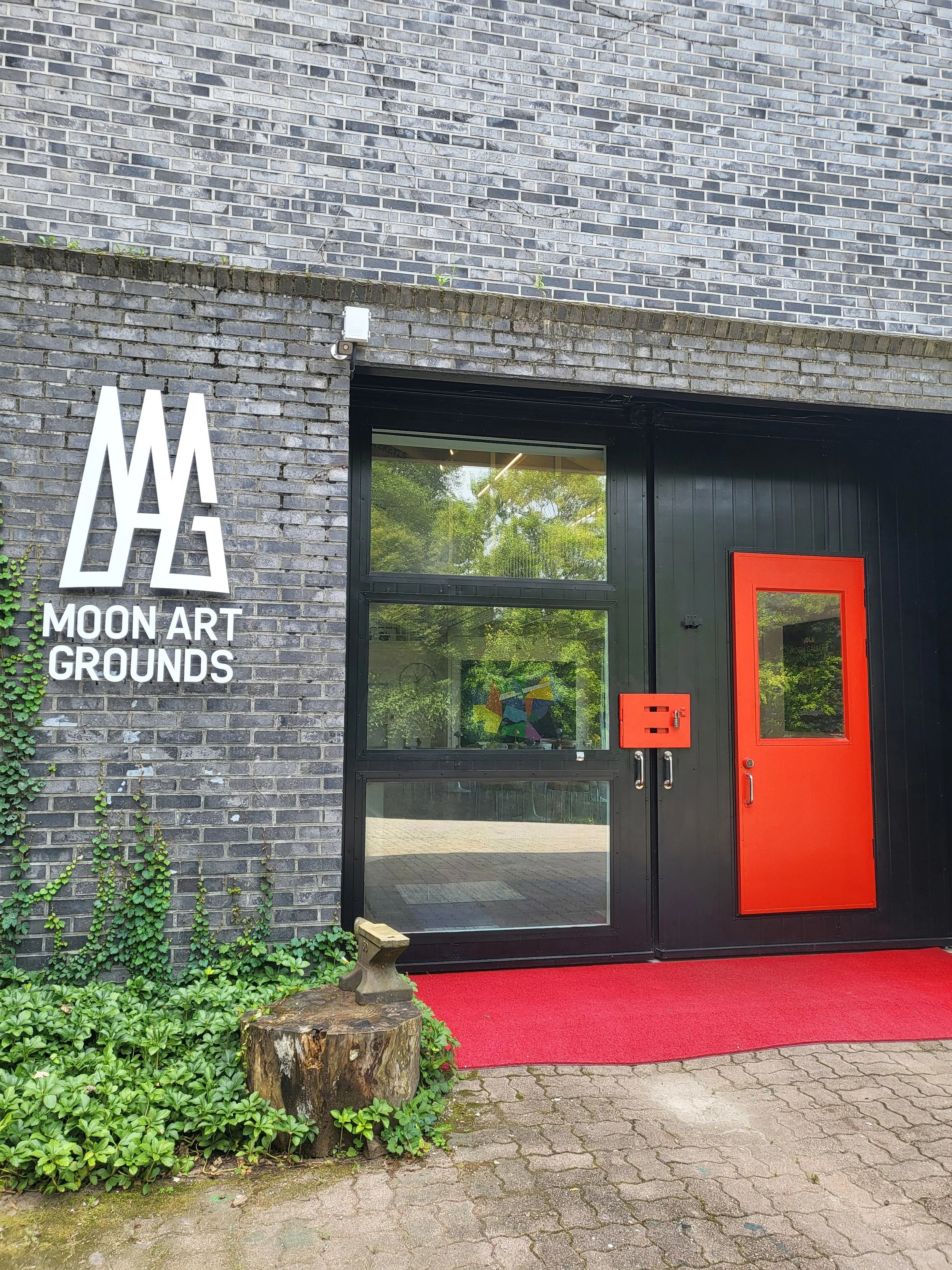 Entrance to Moon Art Grounds gallery — venue hosting Burnsoo Song exhibitions, black brick facade with orange door