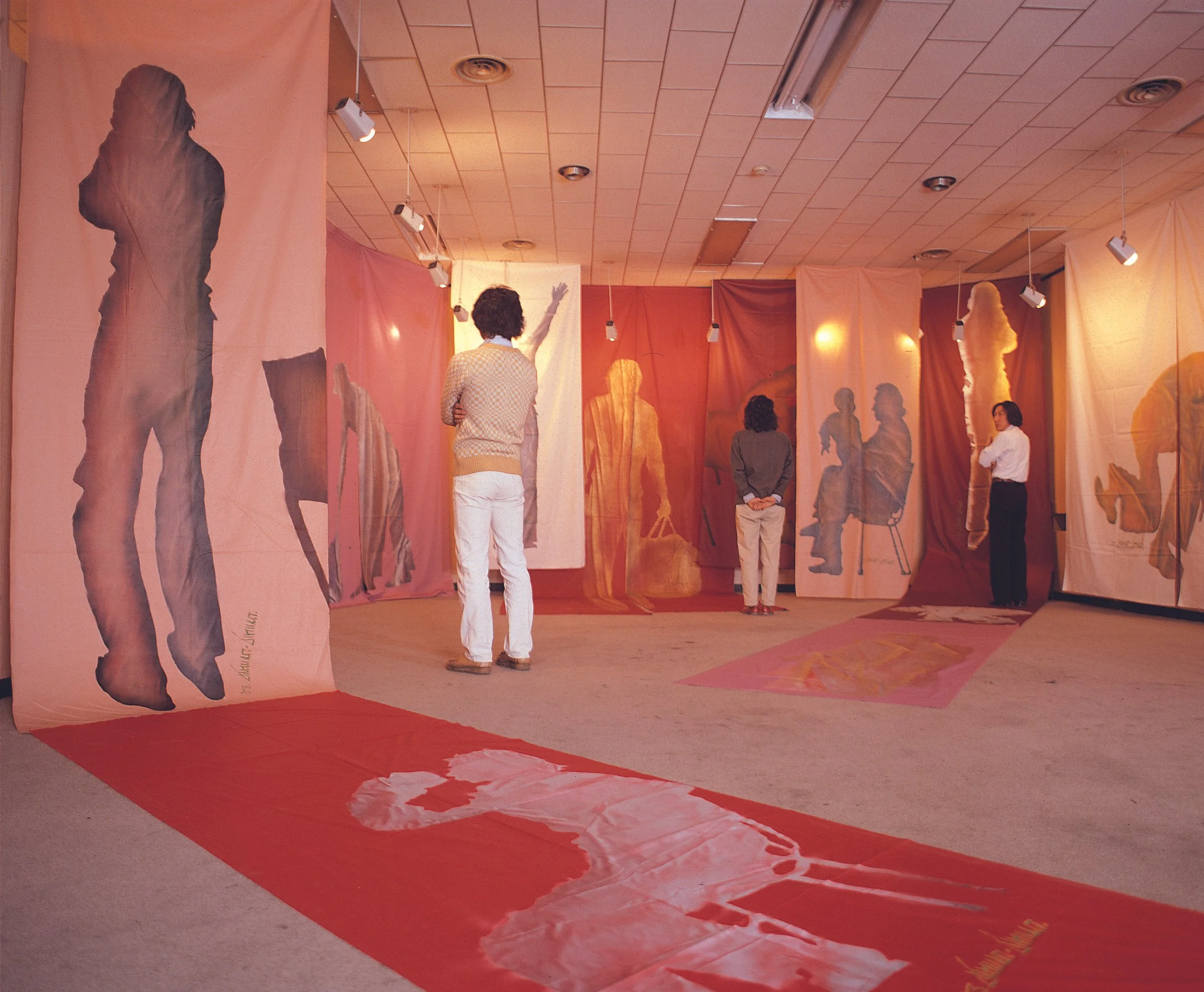 Gallery installation by Burnsoo Song with orange and pink draped silhouette cutouts, viewers engaging with work