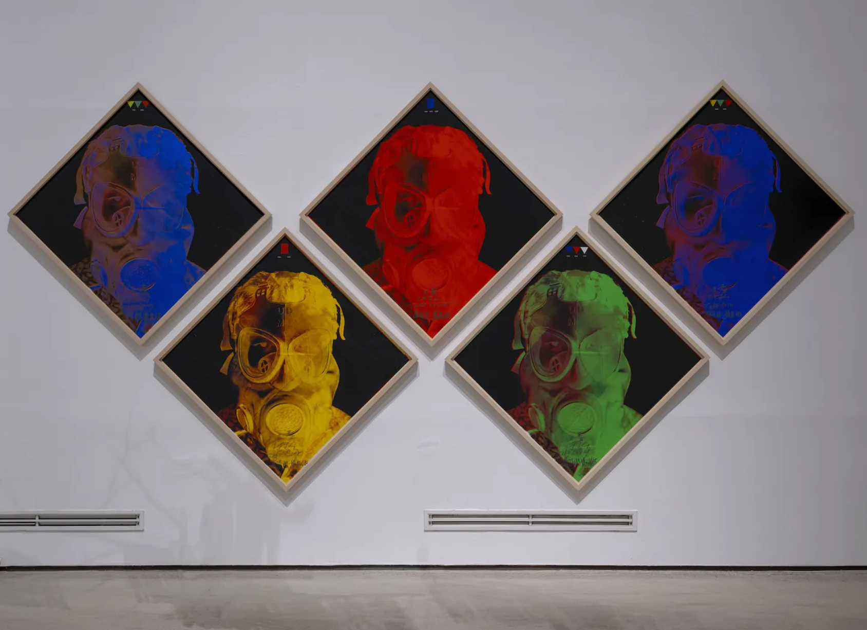 Six framed artworks arranged in a diamond pattern on a white gallery wall. Each features a portrait of a person wearing glasses and a gas mask, with vibrant colors: blue, red, yellow, green, and purple. The artworks have a dark background and include some handwritten text at the bottom.