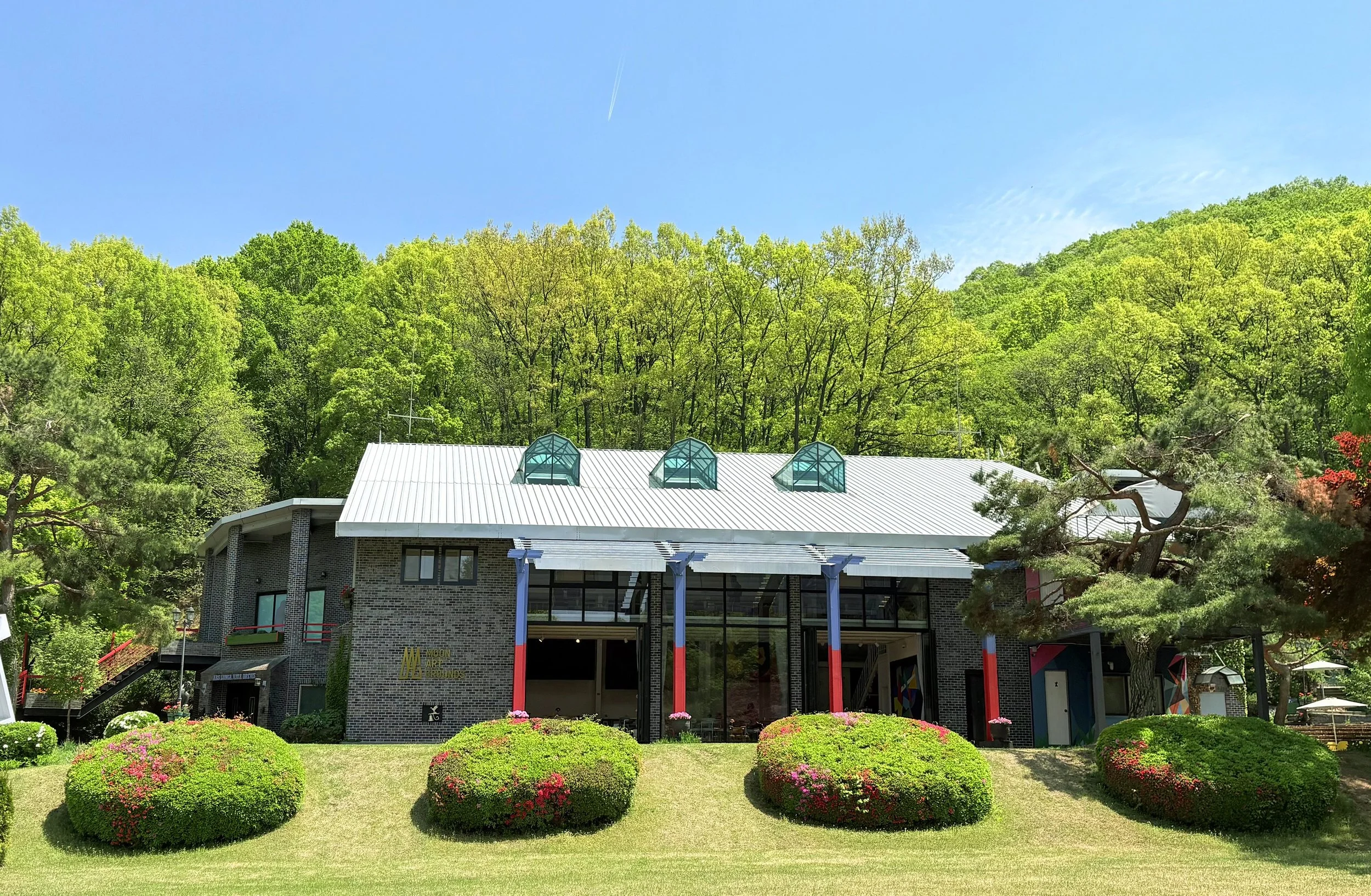 A modern brick building with a white metal roof, three glass dome skylights, and a landscaped foreground with neatly trimmed bushes and colorful flowers, surrounded by tall green trees and a clear blue sky.