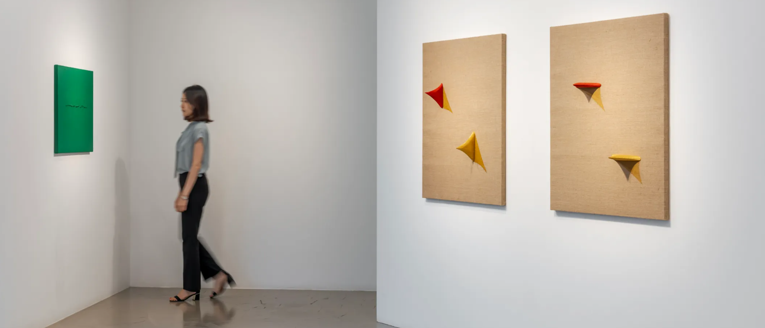 A woman walking in an art gallery with white walls. On the left, a green canvas hangs on the wall, and on the right, two beige panels display small colorful cone-shaped sculptures with shadows.