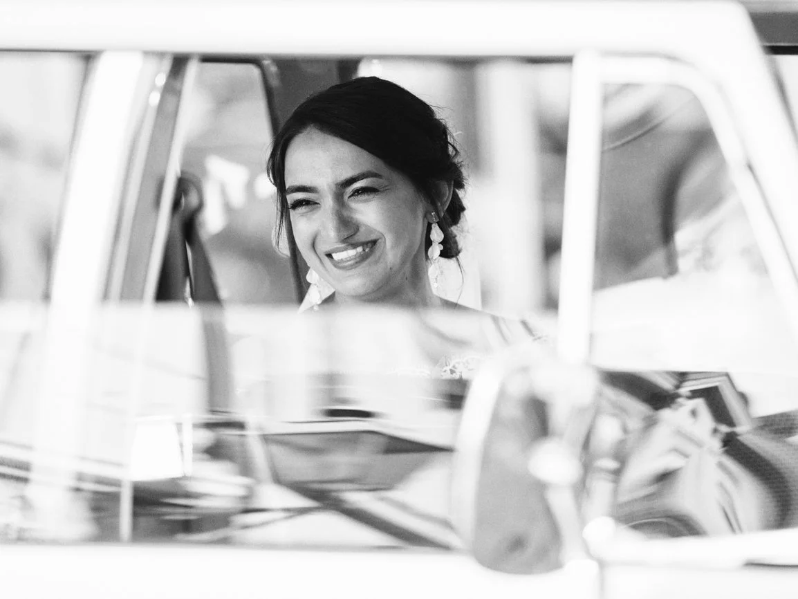 Portrait en noir et blanc d'une mariée souriante photographiée à travers la fenêtre d'une voiture ancienne, capturant un instant spontané et élégant avant la cérémonie de mariage en Suisse.