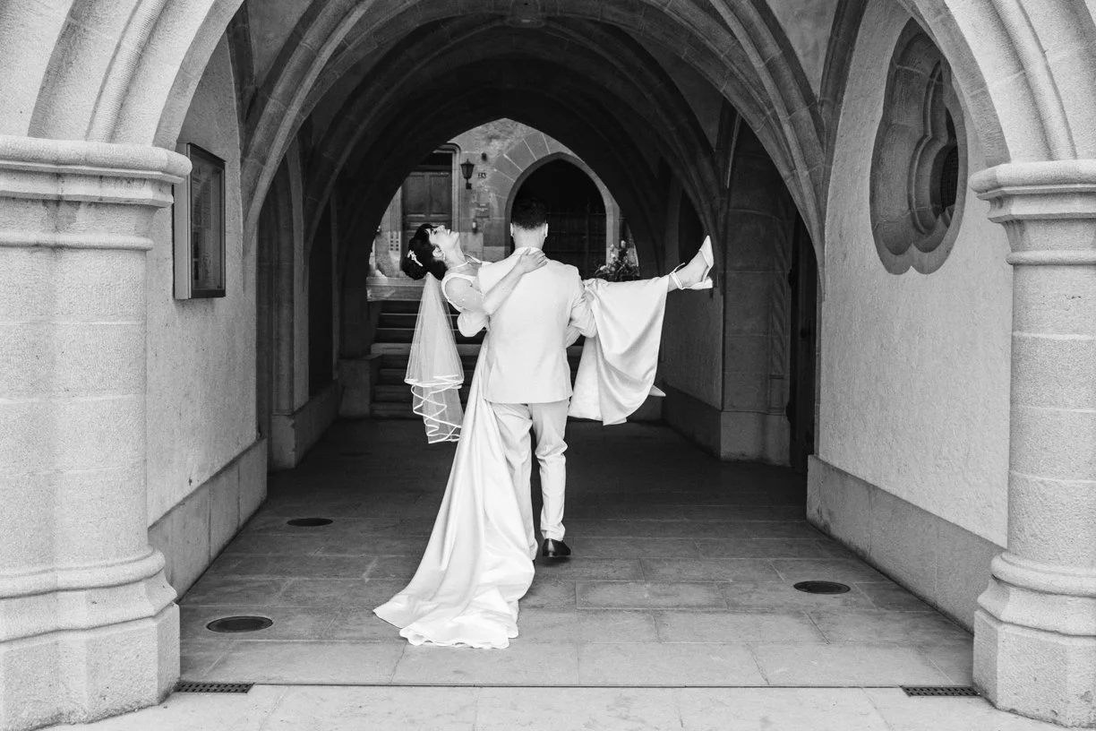 Le marié porte sa mariée sous les arches d'un bâtiment historique dans une scène romantique en noir et blanc, capturant un instant de complicité lors d'un mariage en Suisse