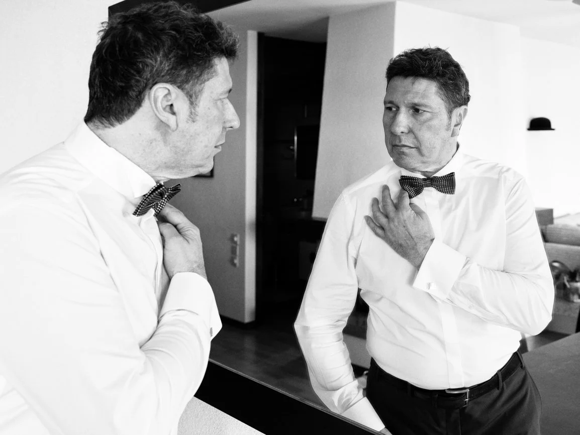 Le marié ajuste son nœud papillon devant un miroir pendant les préparatifs, photographie de mariage en noir et blanc capturant un instant élégant et authentique avant la cérémonie en Suisse